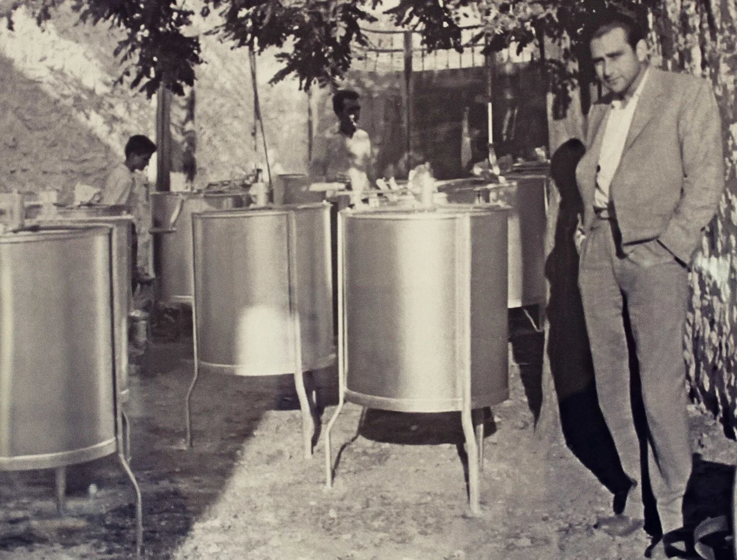 Black and white photograph of a man in a suit standing outdoors beside a row of cylindrical metal tables, with children and a woman in the background under a tree canopy, possibly at an outdoor event or fair.