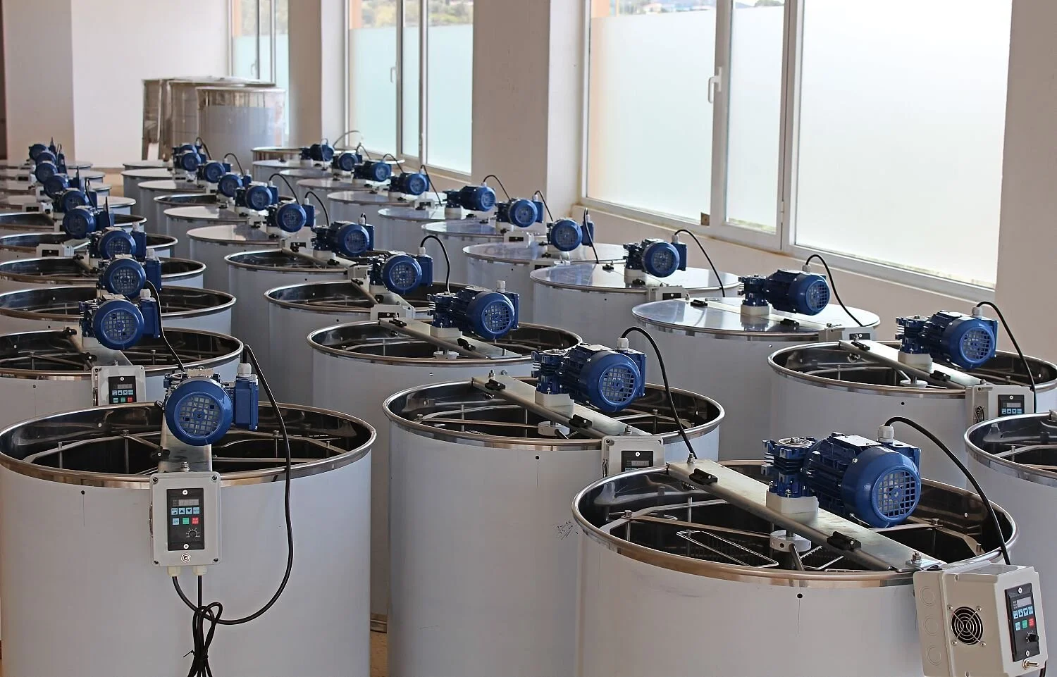 Multiple large white industrial machines with blue motors and control panels inside a room with windows.