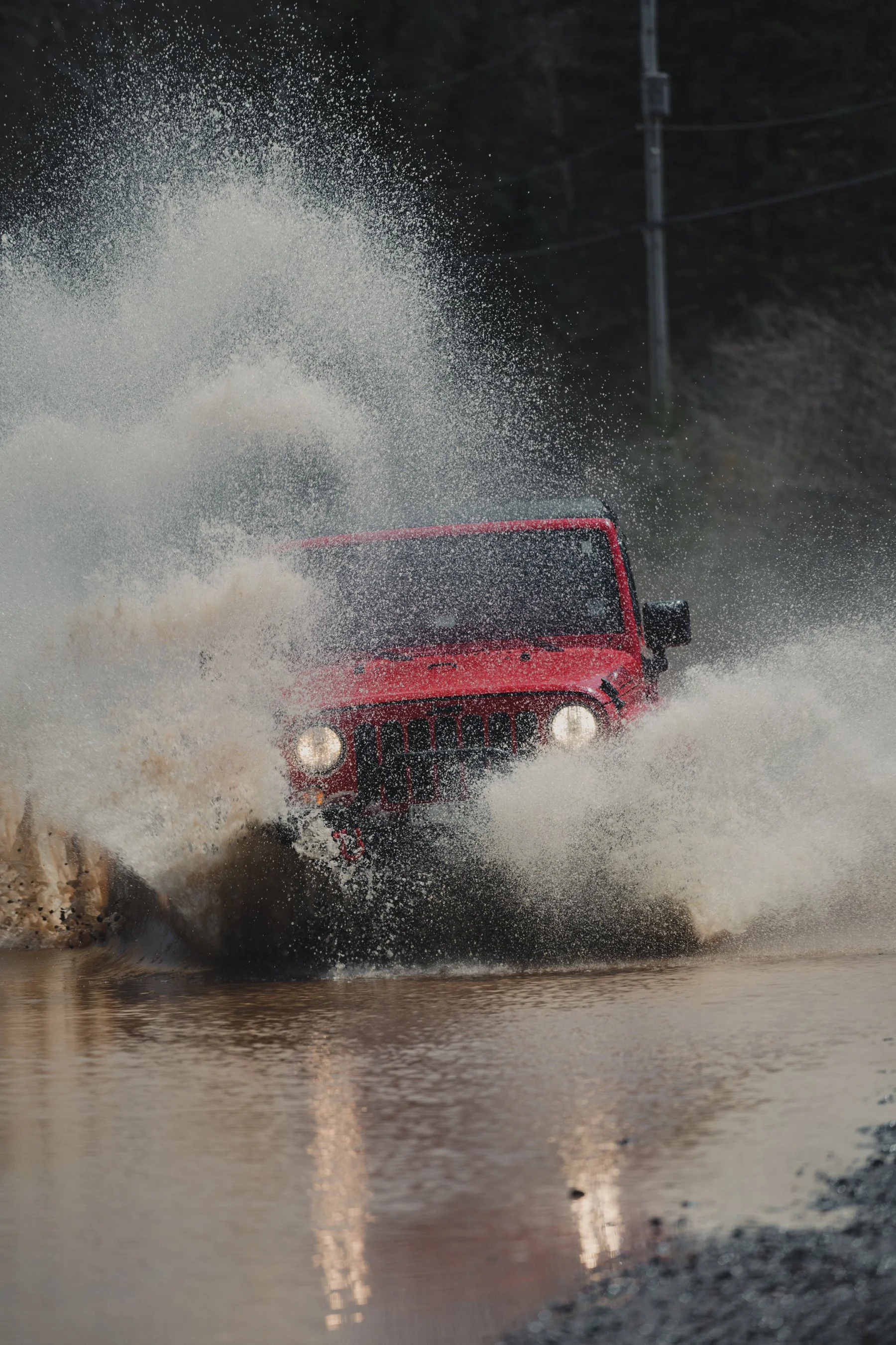 A red Jeep driving through a muddy water crossing, creating a splash.