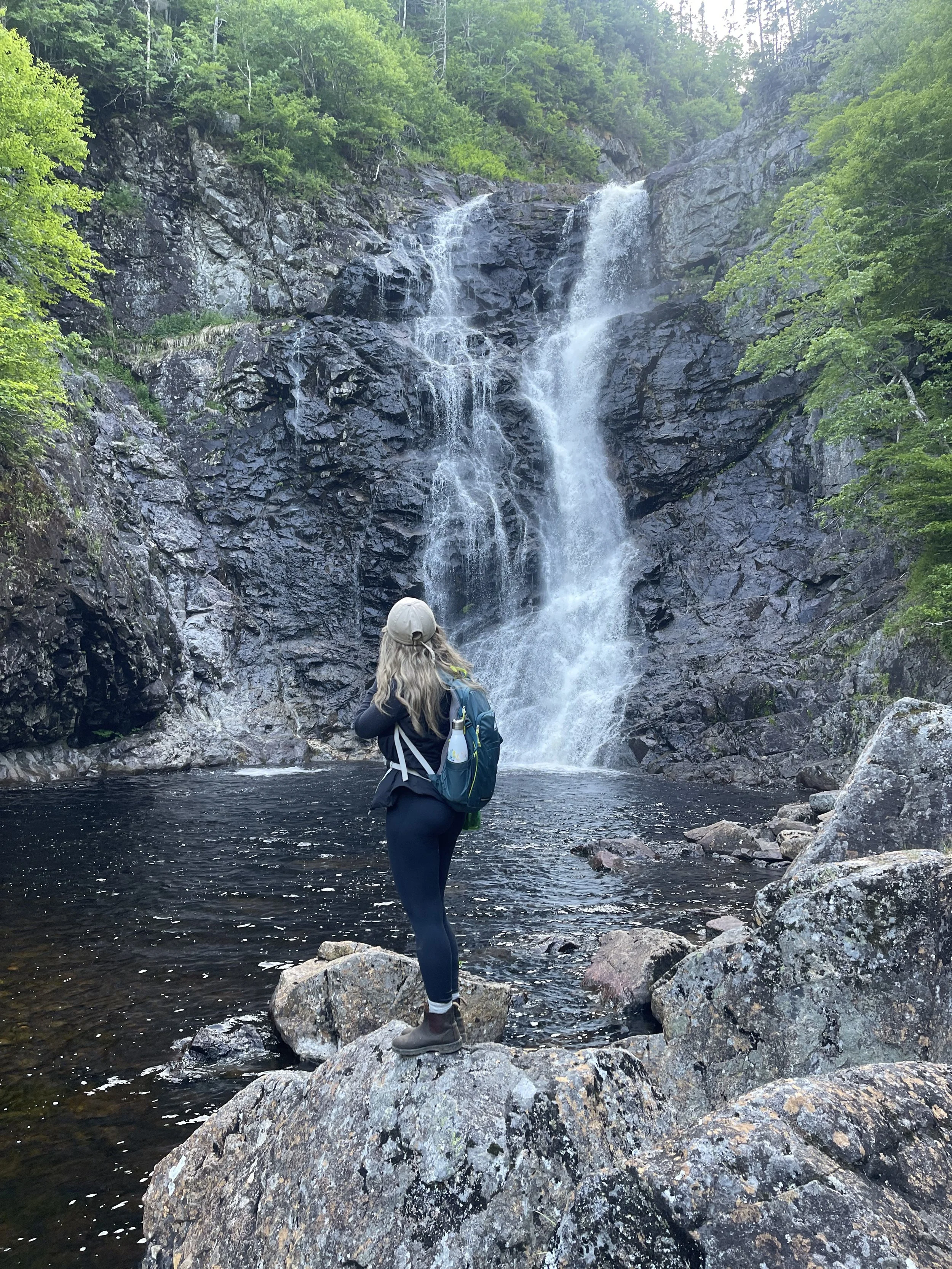 A woman with long hair, wearing a tan cap, dark backpack, dark jacket, and dark pants, standing on a large rock in front of a waterfall in a forested area.