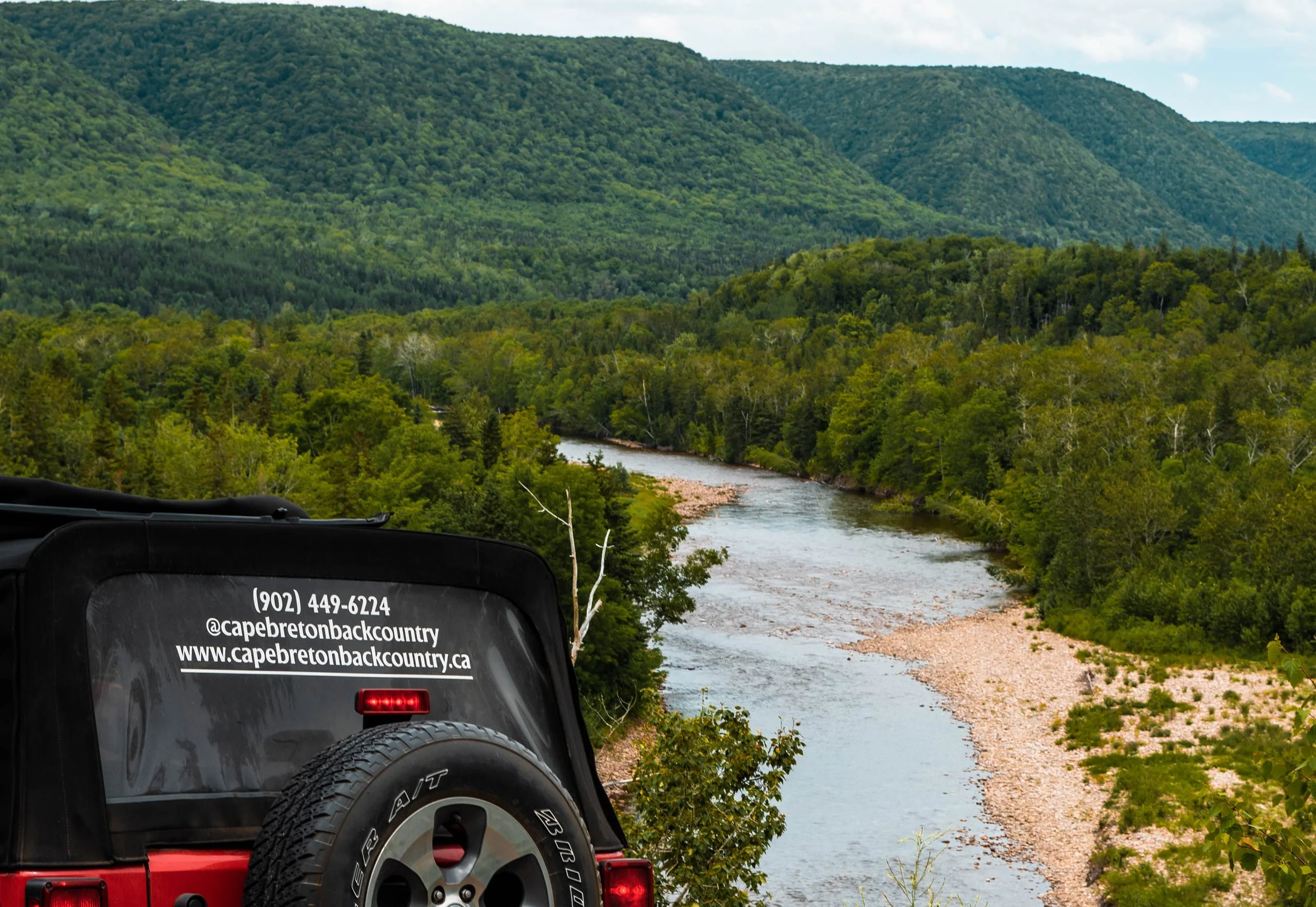 Back of a red Jeep with Cape Breton Backcountry Adventures branding overlooking the Margaree River valley with dense forest and mountains.