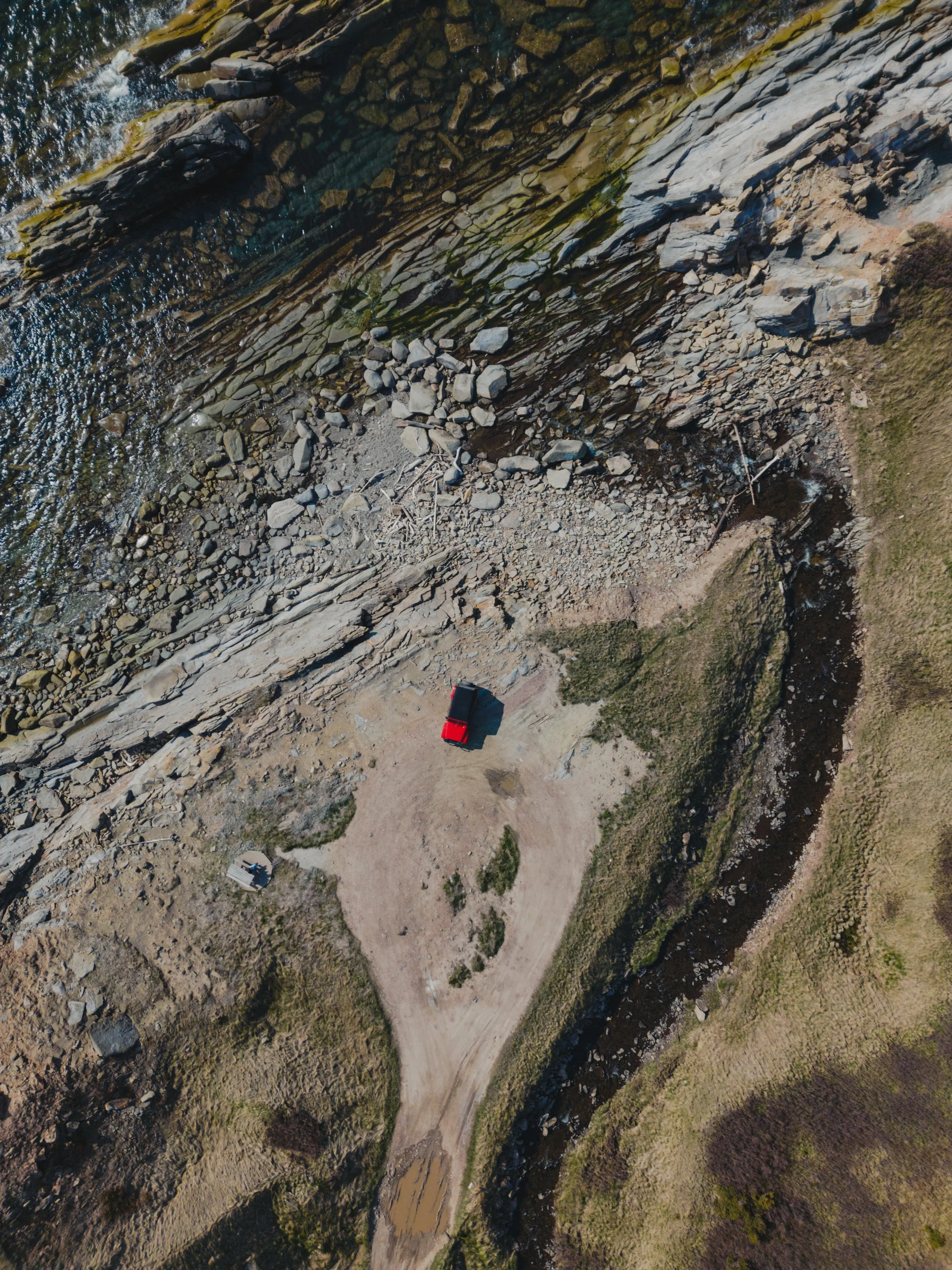 Aerial view of a rocky shoreline with a small stream running through grassy terrain, and a red vehicle parked on a dirt area near the water.