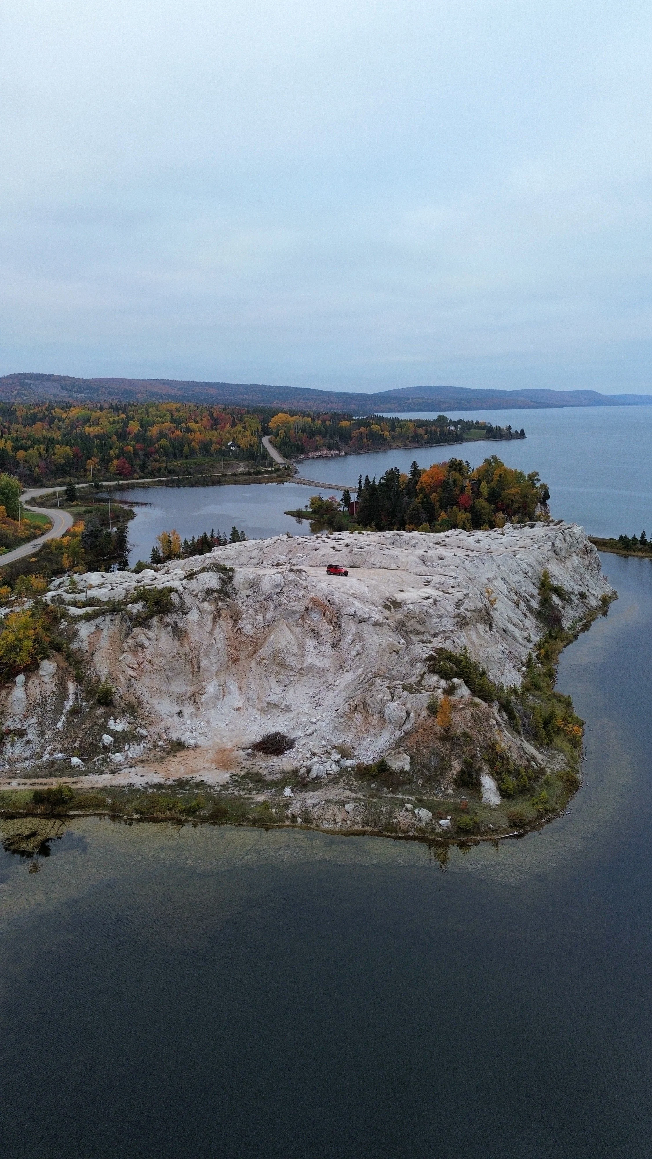 Aerial view of a rocky hill surrounded by water, with a red Jeep parked on top and trees at the base, during autumn with colorful foliage.