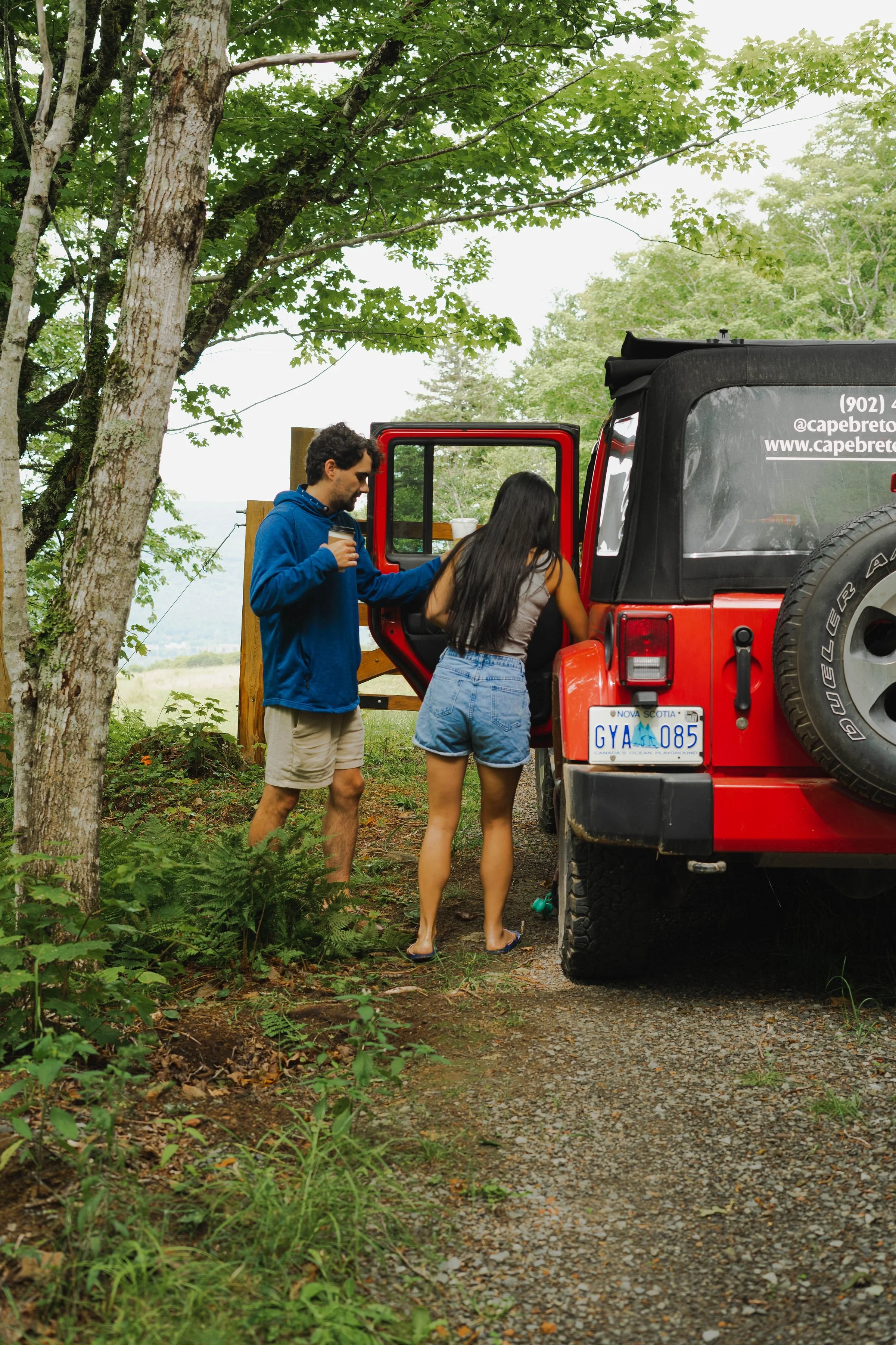 Two people near a red Jeep parked on a dirt trail next to trees in a forested area, with one person entering the vehicle and the other holding an iced coffee, during daytime.