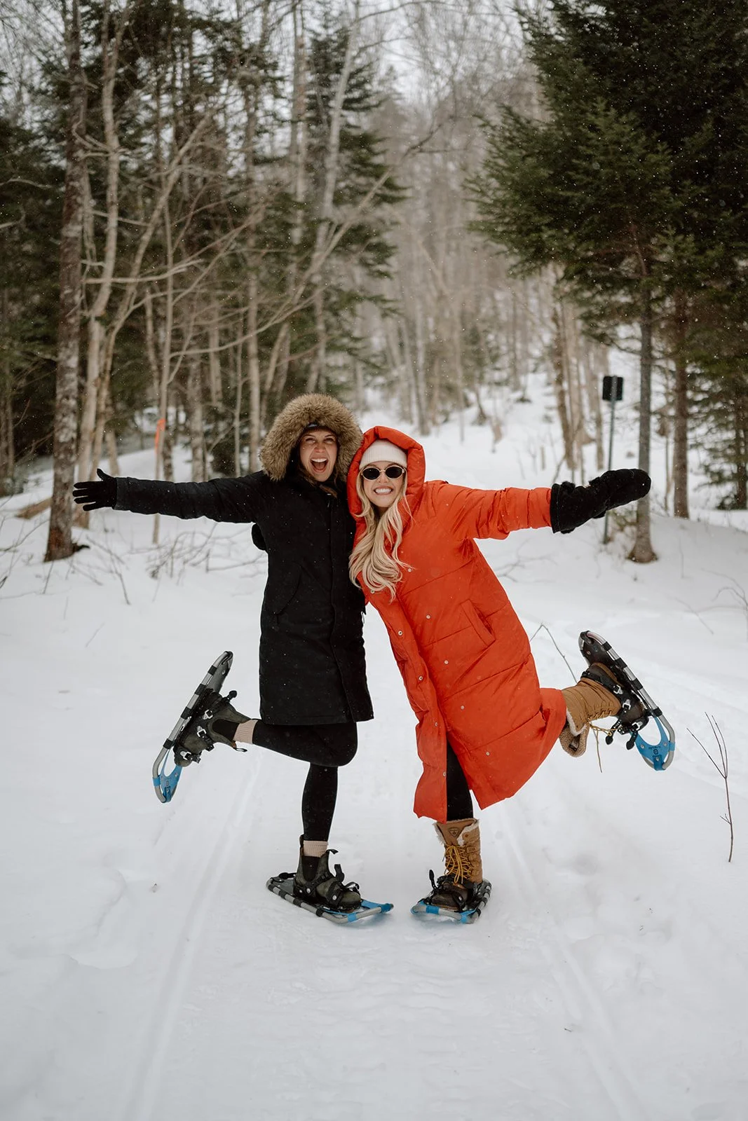 Two women in black and orange winter coats snowboarding and posing with arms outstretched in a snowy forest during a Maple Adventure Tour in Cape Breton.