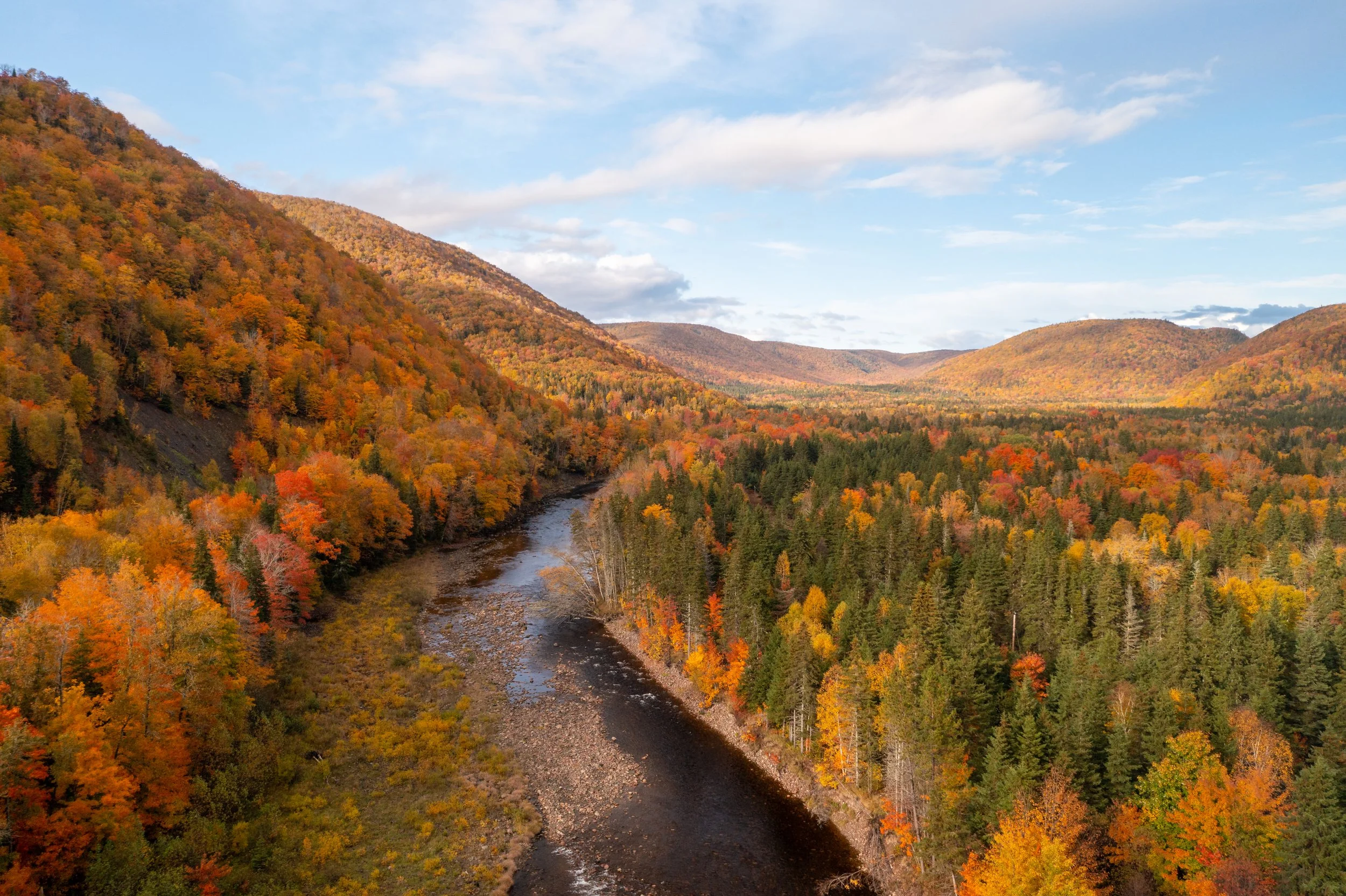 An aerial view of a river winding through a forested valley with colorful autumn foliage and rolling hills in the background under a partly cloudy sky.