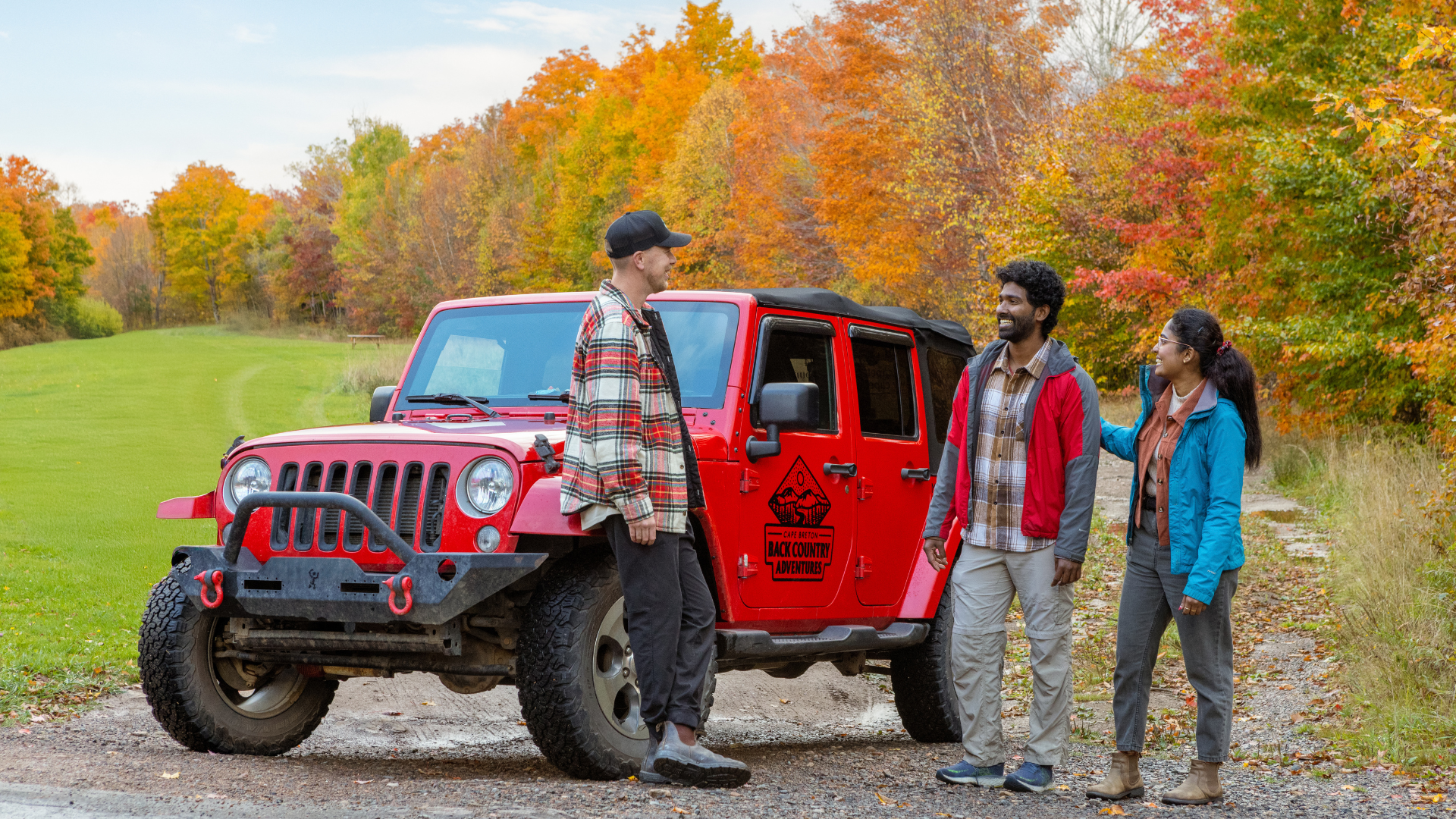 Three people smiling and talking near a red Jeep with a 'Cape Breton Back Country Adventures' logo,  surrounded by autumn-colored trees in a park.