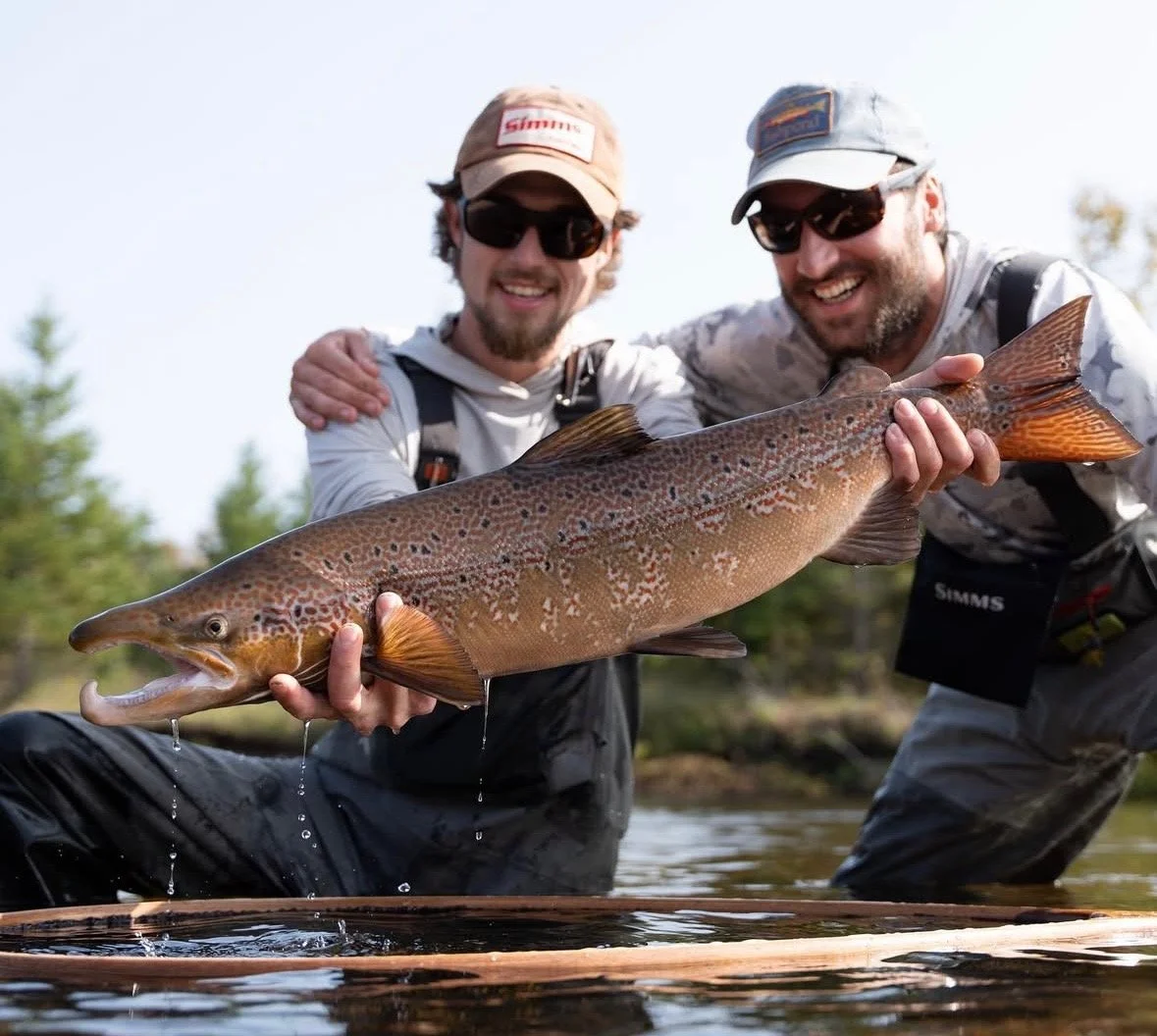 Two men wearing sunglasses and fishing gear holding a large fish near a body of water, smiling.
