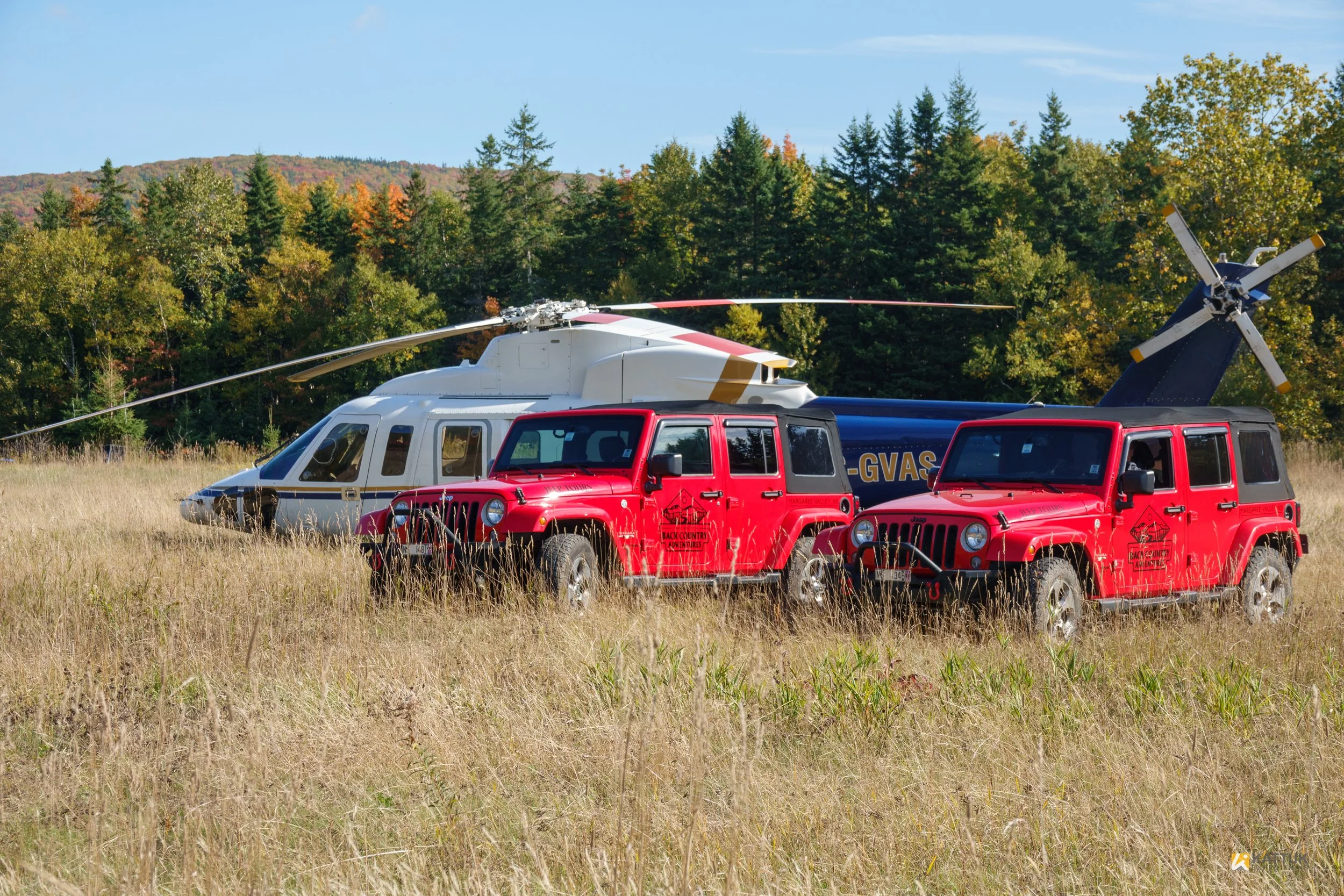 Two red Jeep Wranglers and a gray helicopter with a blue tail parked in a grassy field with a tree-lined mountain in the background.