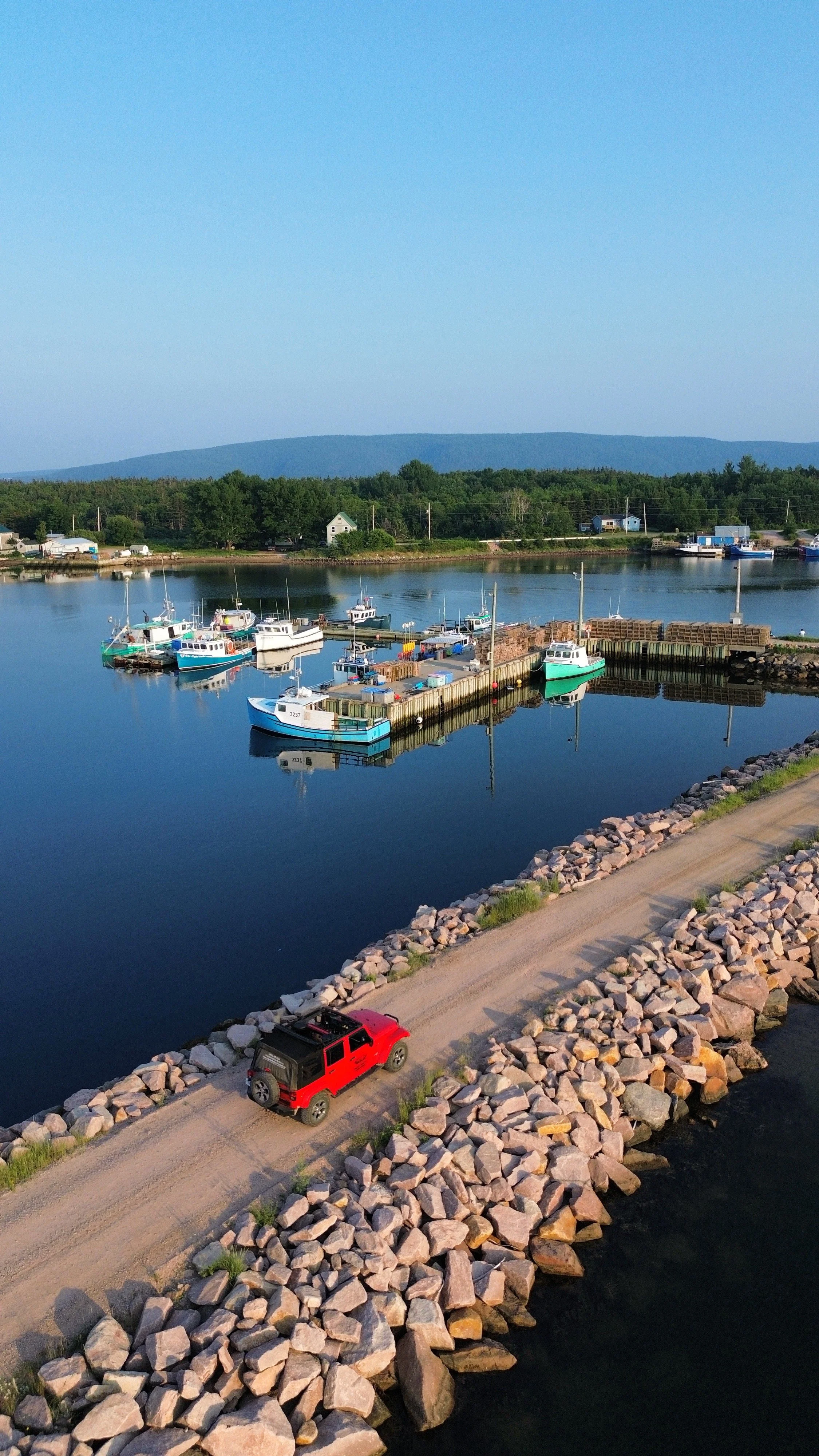 Aerial view of a small harbor with boats docked at the pier, a red Jeep driving along a rocky road beside the water, green trees, houses, and distant hills under a clear blue sky.