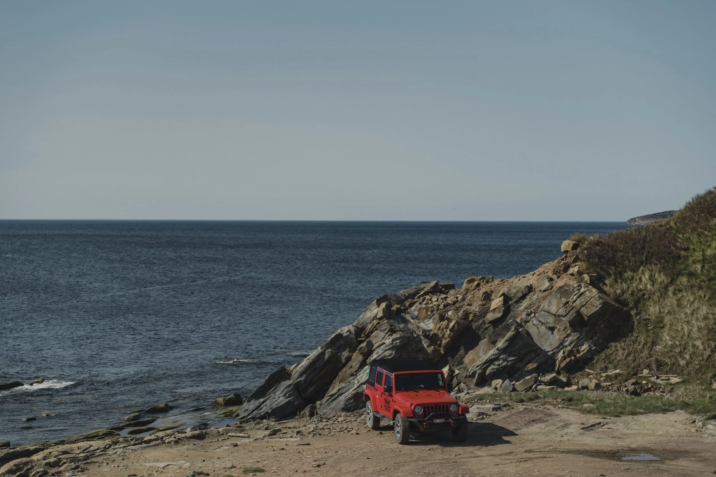 Red off-road Jeep parked on a rocky beach with the ocean in the background.
