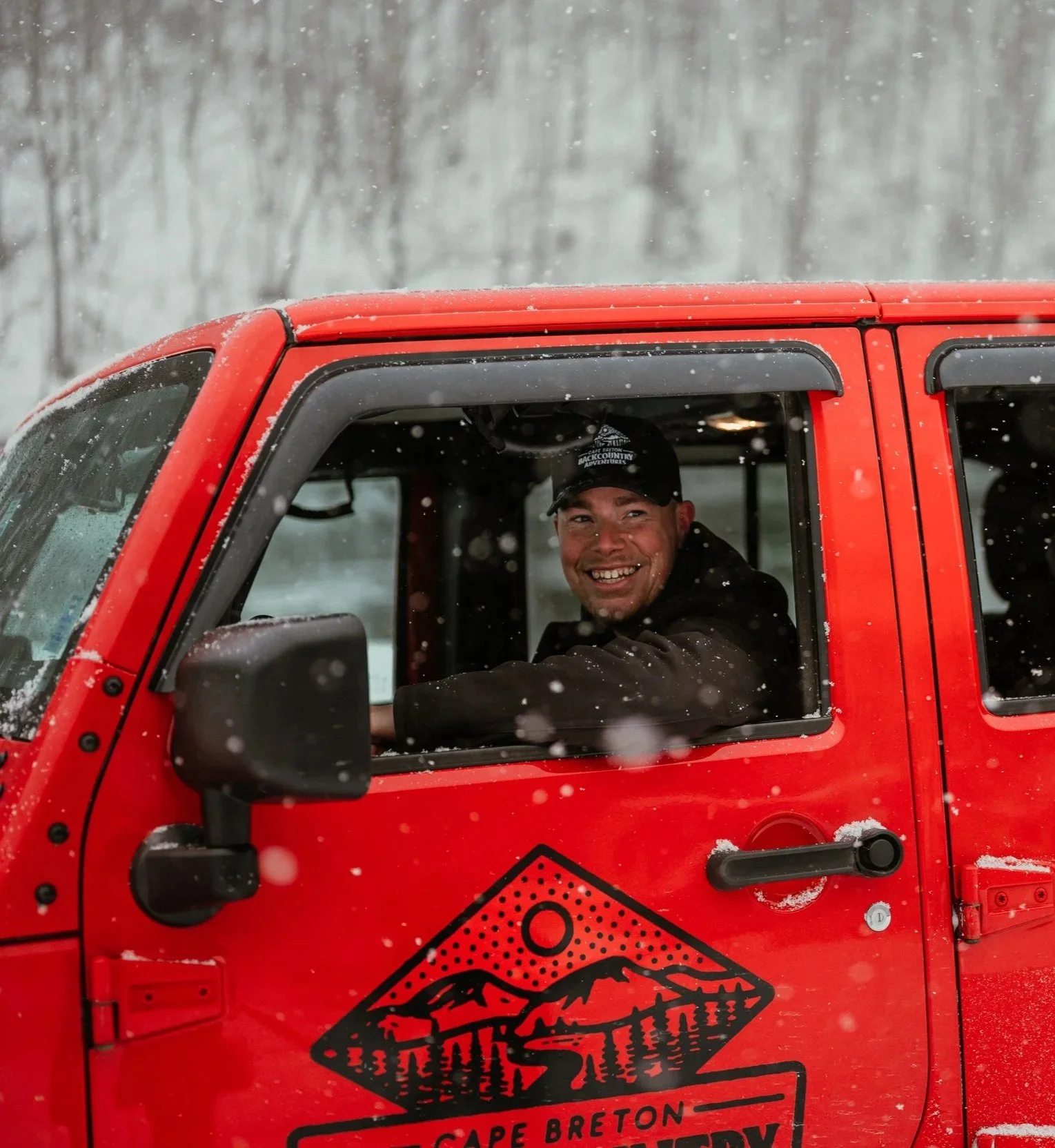A smiling man sitting in the driver's seat of a red vehicle with a logo that reads 'Cape Breton Back Country Adventures' and features a mountain, trees, and a river. Snow is falling.