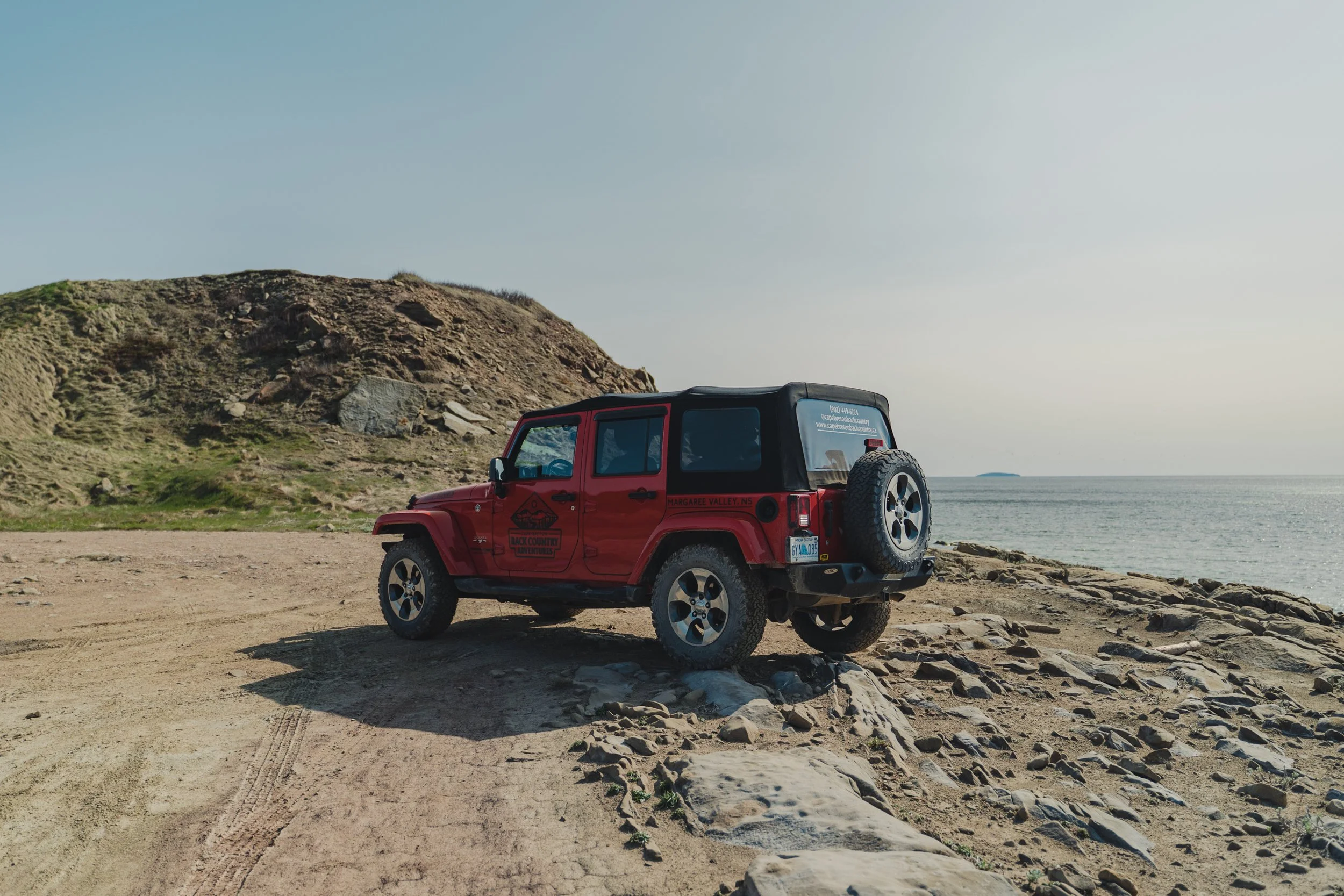 A red Jeep with a 'Cape Breton Back Country Adventures' logo parked on a coastal trail overlooking the ocean and rocky shoreline in Cape Breton.