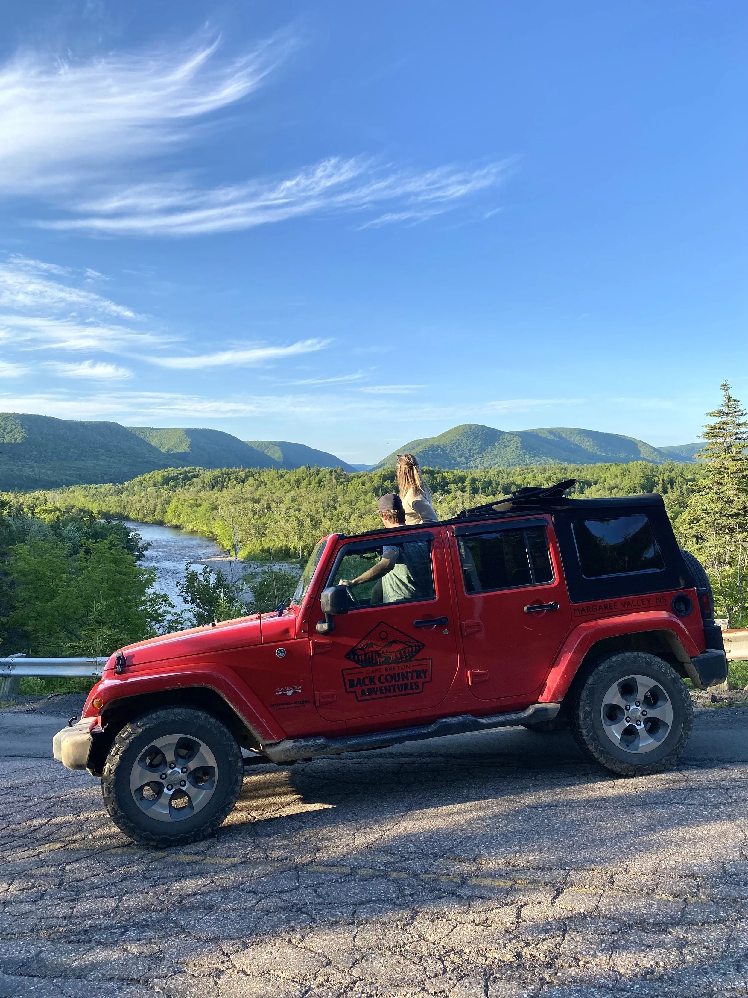 A red Jeep parked on a cracked asphalt road overlooking a river and lush green forest with mountains in the background under a partly cloudy blue sky. Two people are inside and one is standing through the open roof.