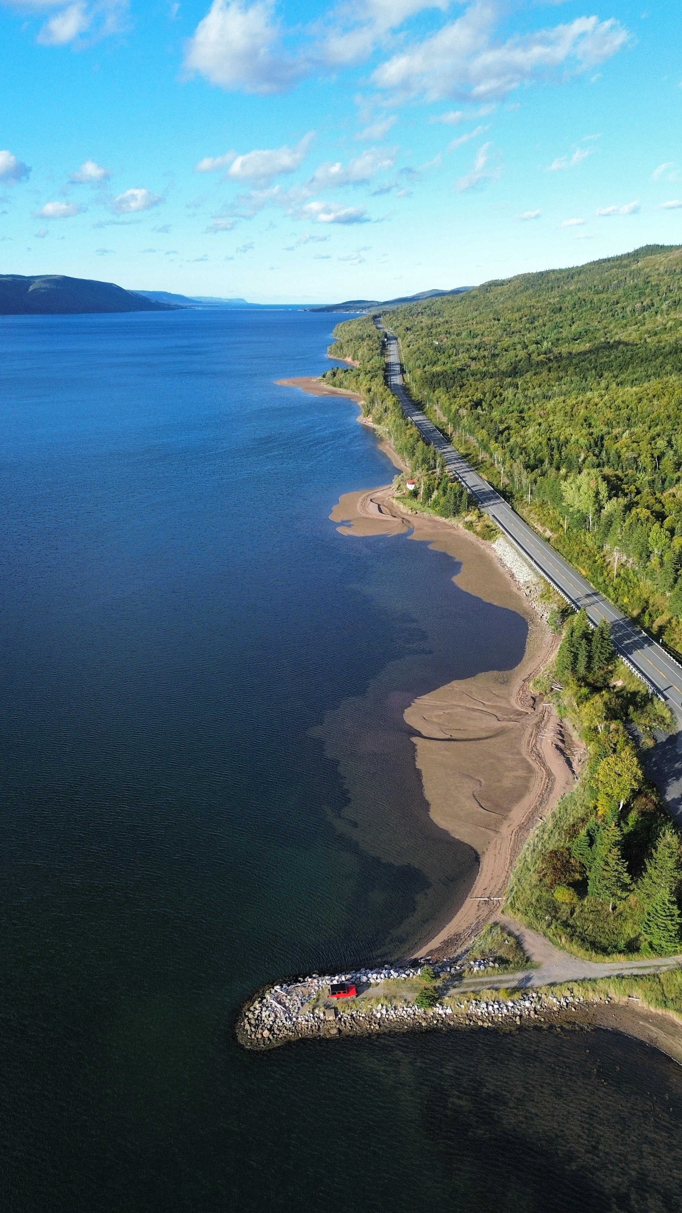 An aerial view of a large body of water next to a forested shoreline. A road runs parallel to the water, with a small dock and a red buoy near the shore. The sky is partly cloudy.