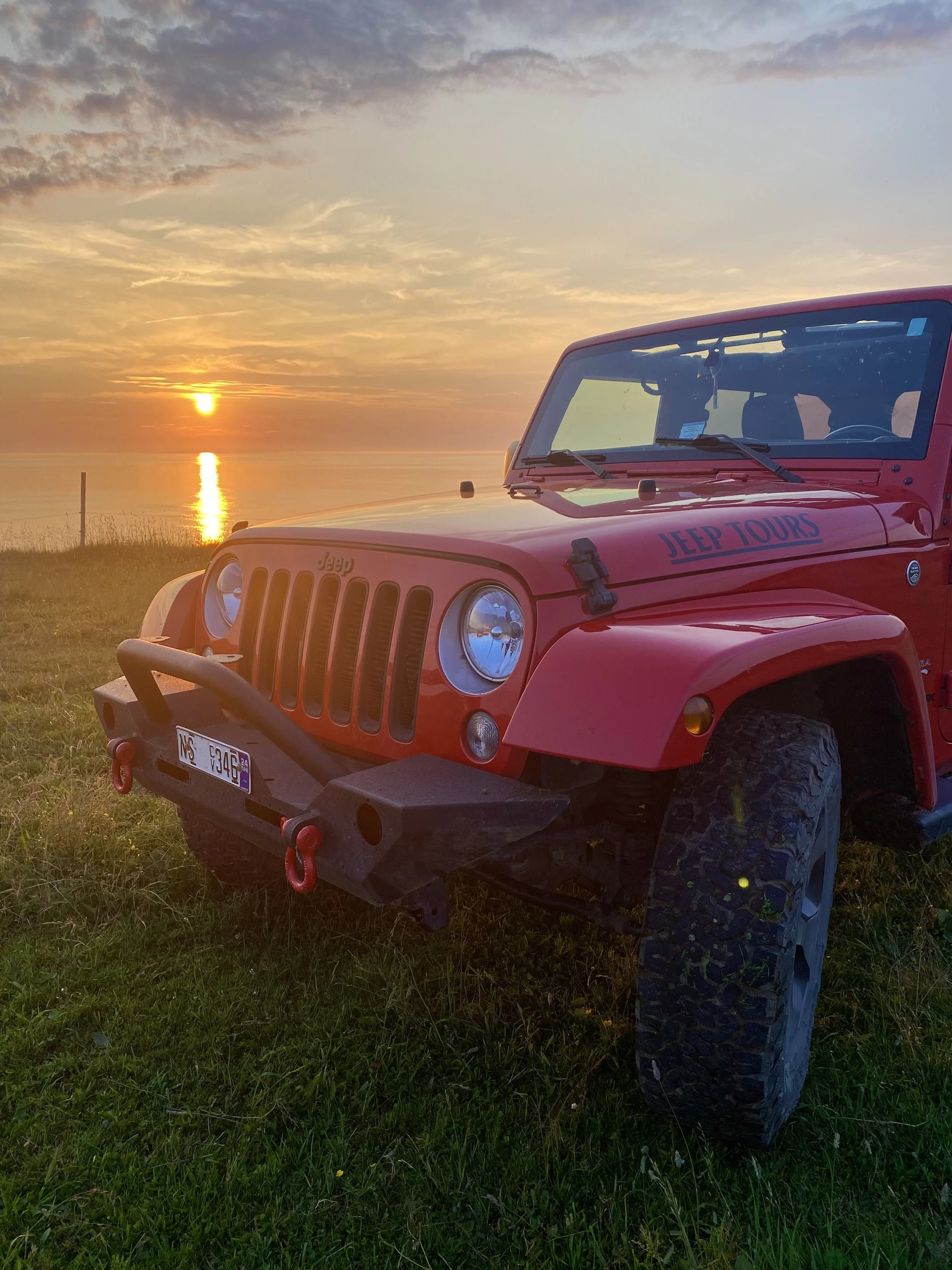 Red Jeep parked on a grassy coastal overlook in Cape Breton, with the sun setting over the ocean and golden light reflecting across the water.