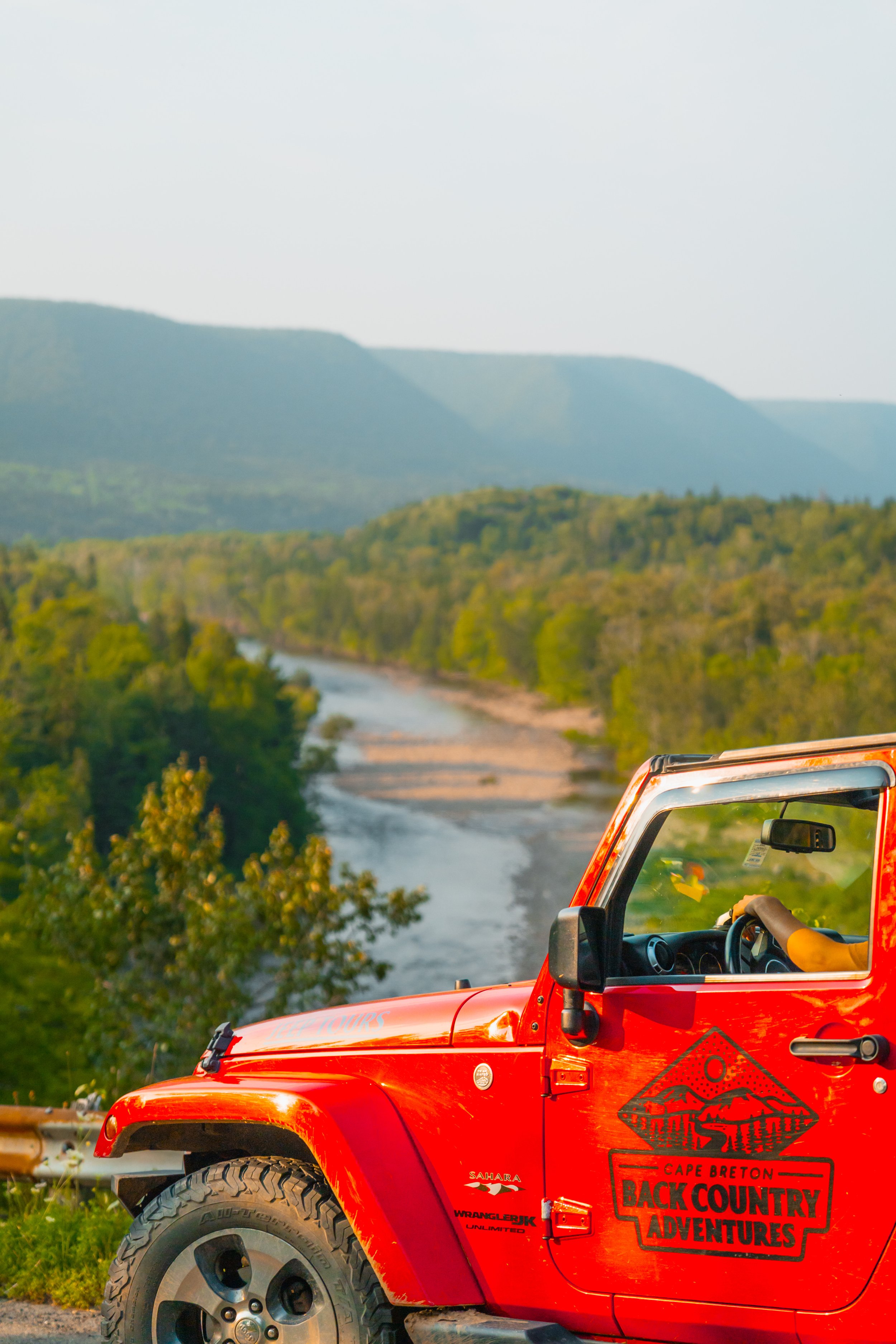 Red Cape Breton Backcountry Adventures Jeep overlooking the Margaree River valley, with forested hills and mountains in the background on a scenic summer day.
