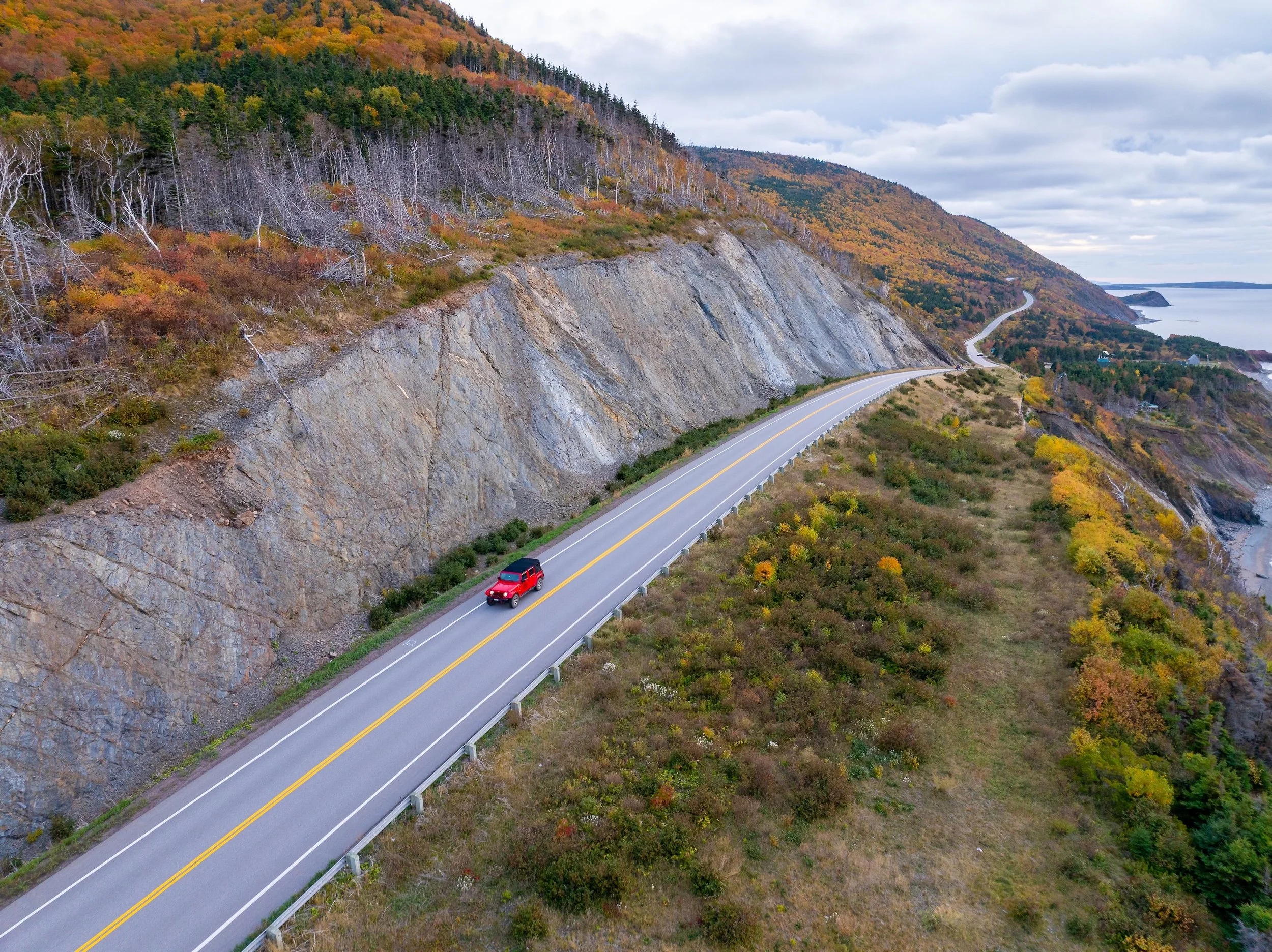 Red Jeep driving along a winding coastal section of the Cabot Trail in Cape Breton, surrounded by rugged cliffs and vibrant fall foliage overlooking the ocean.