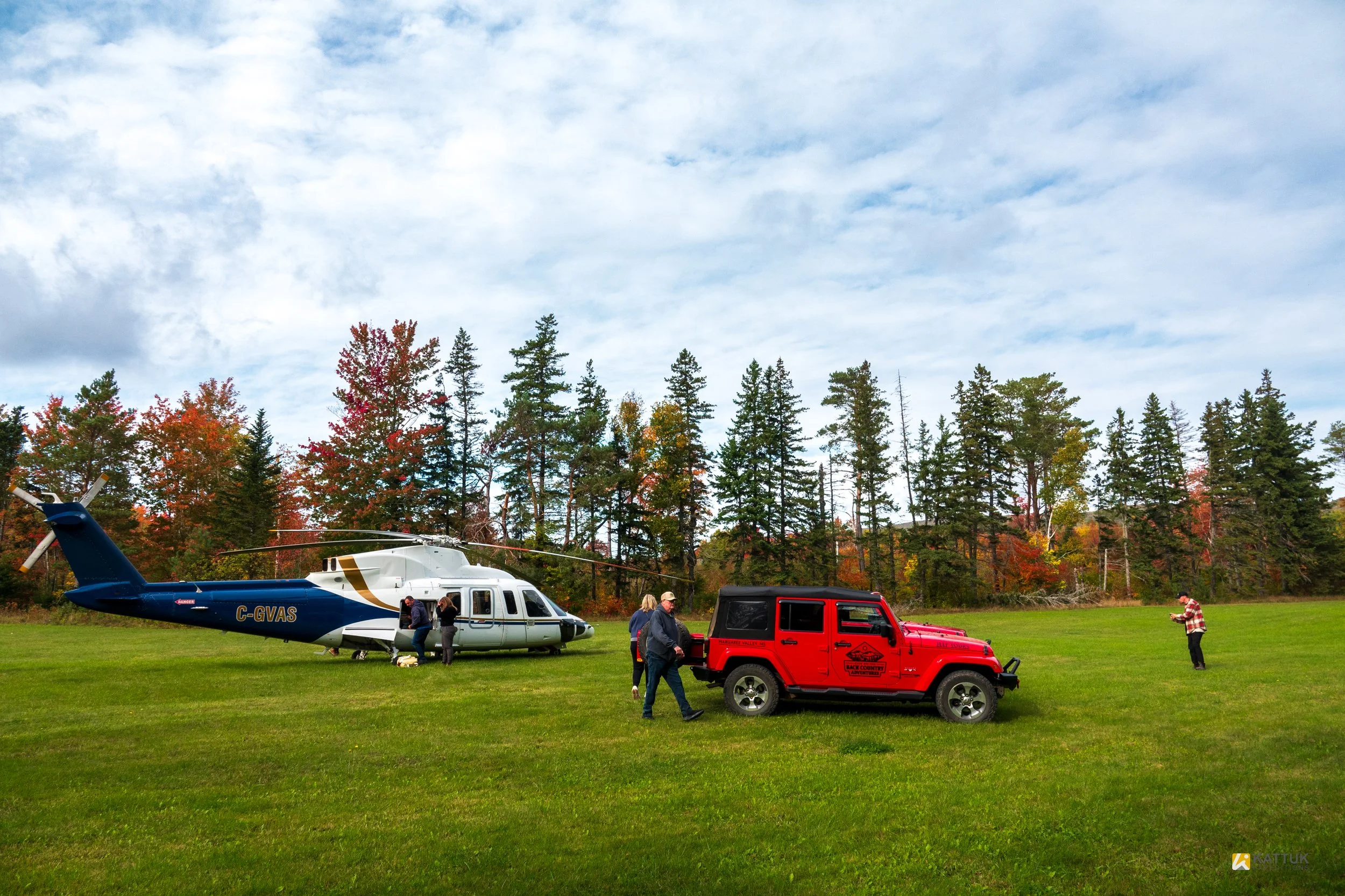 People gathered around a helicopter and a red Jeep in a grassy field with a backdrop of colorful autumn trees and a partly cloudy sky.