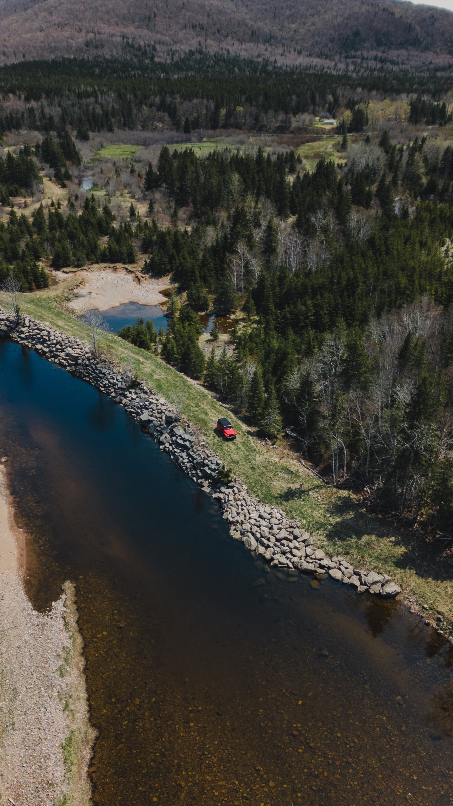 Aerial view of a landscape with a river bordered by rocks, surrounded by grassy areas and dense forest, with a red Jeep parked near the riverbank.