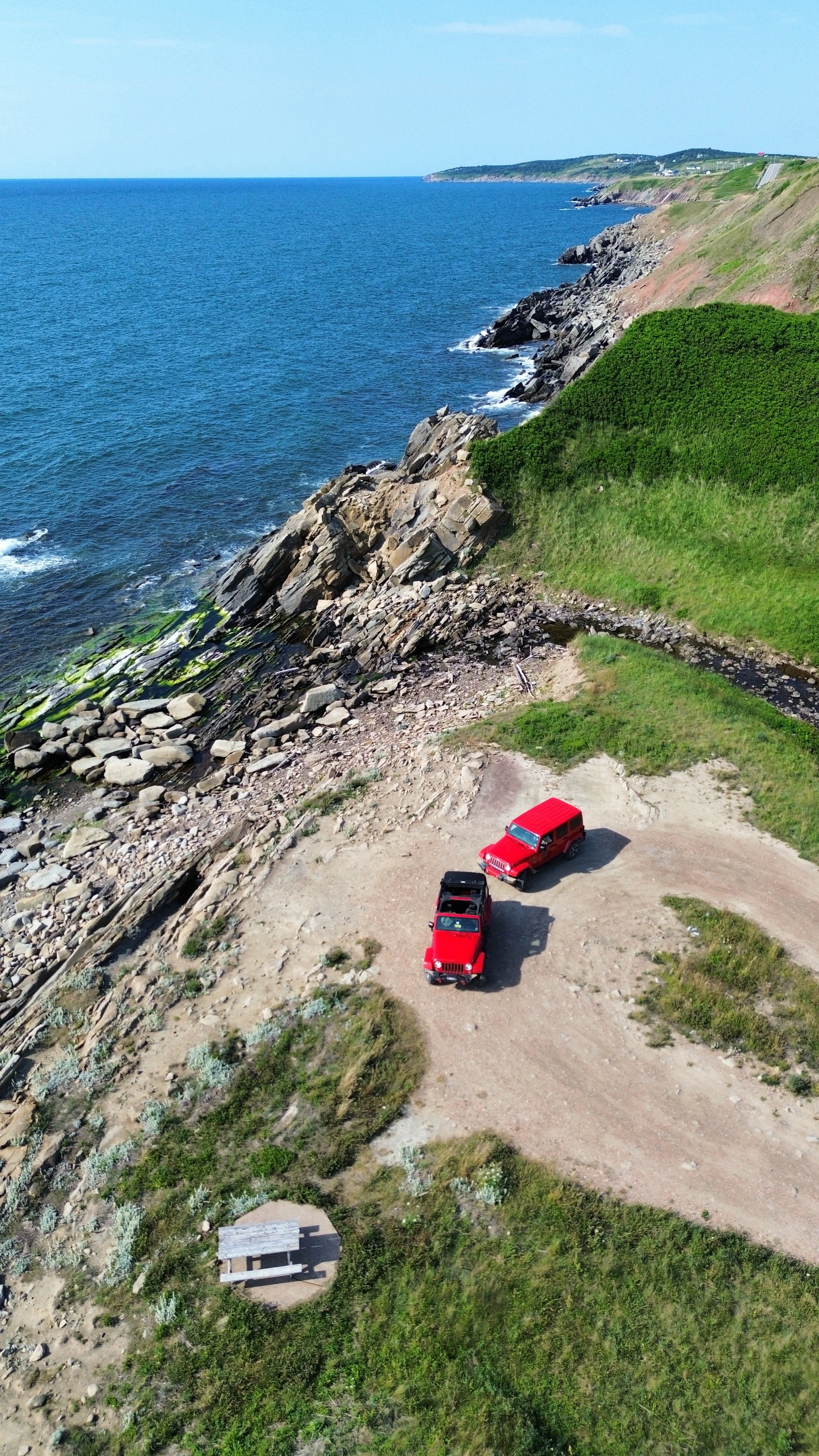 An aerial view of two red Jeep Wranglers parked on a dirt clearing near a rocky coastline with waves and green hills in the background.