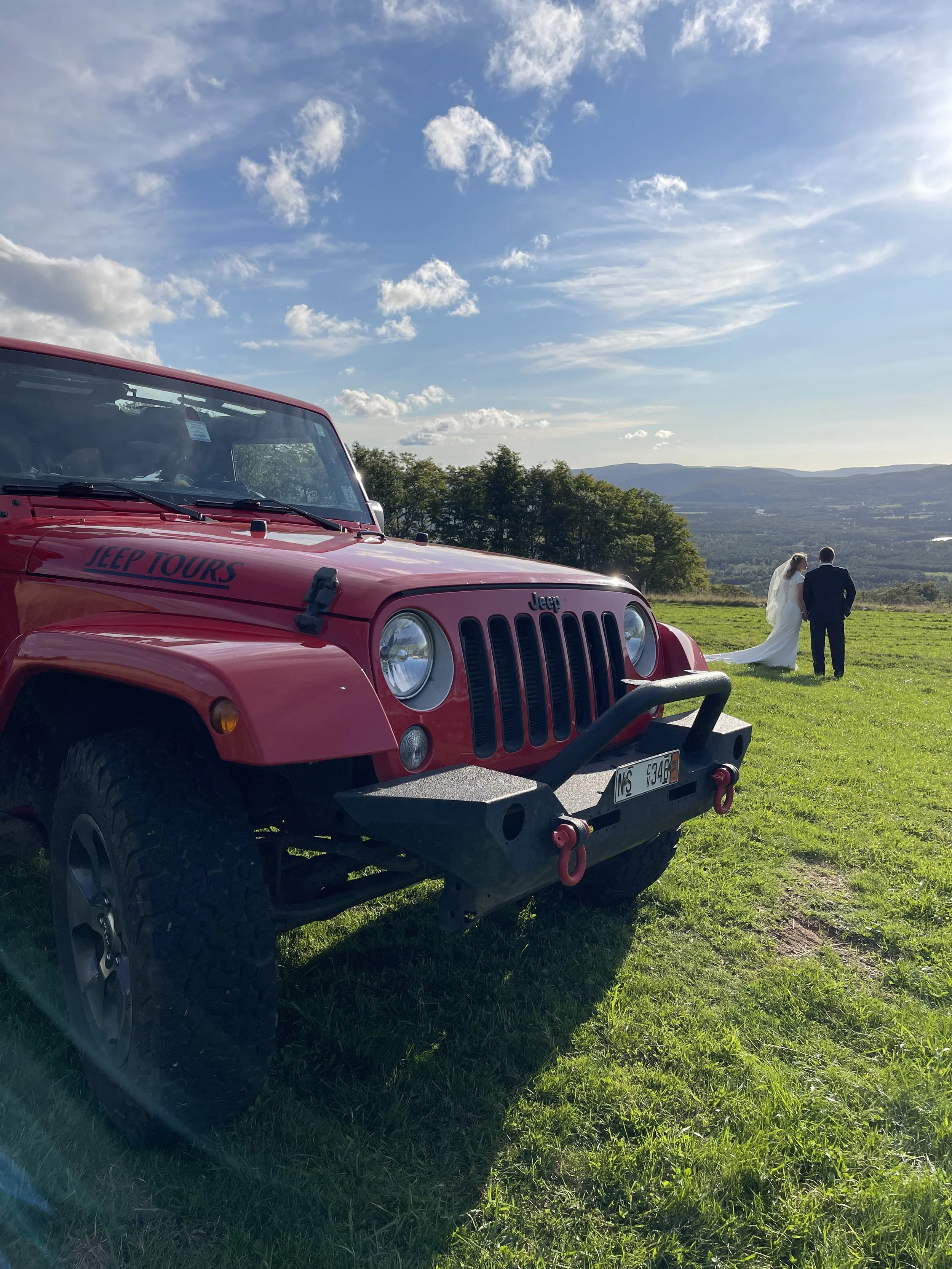 A red Jeep with 'Jeep Tours' written on the hood parked on a grassy hill with a view of trees, mountains, and a clear blue sky. In the background, a bride and groom are walking hand-in-hand.