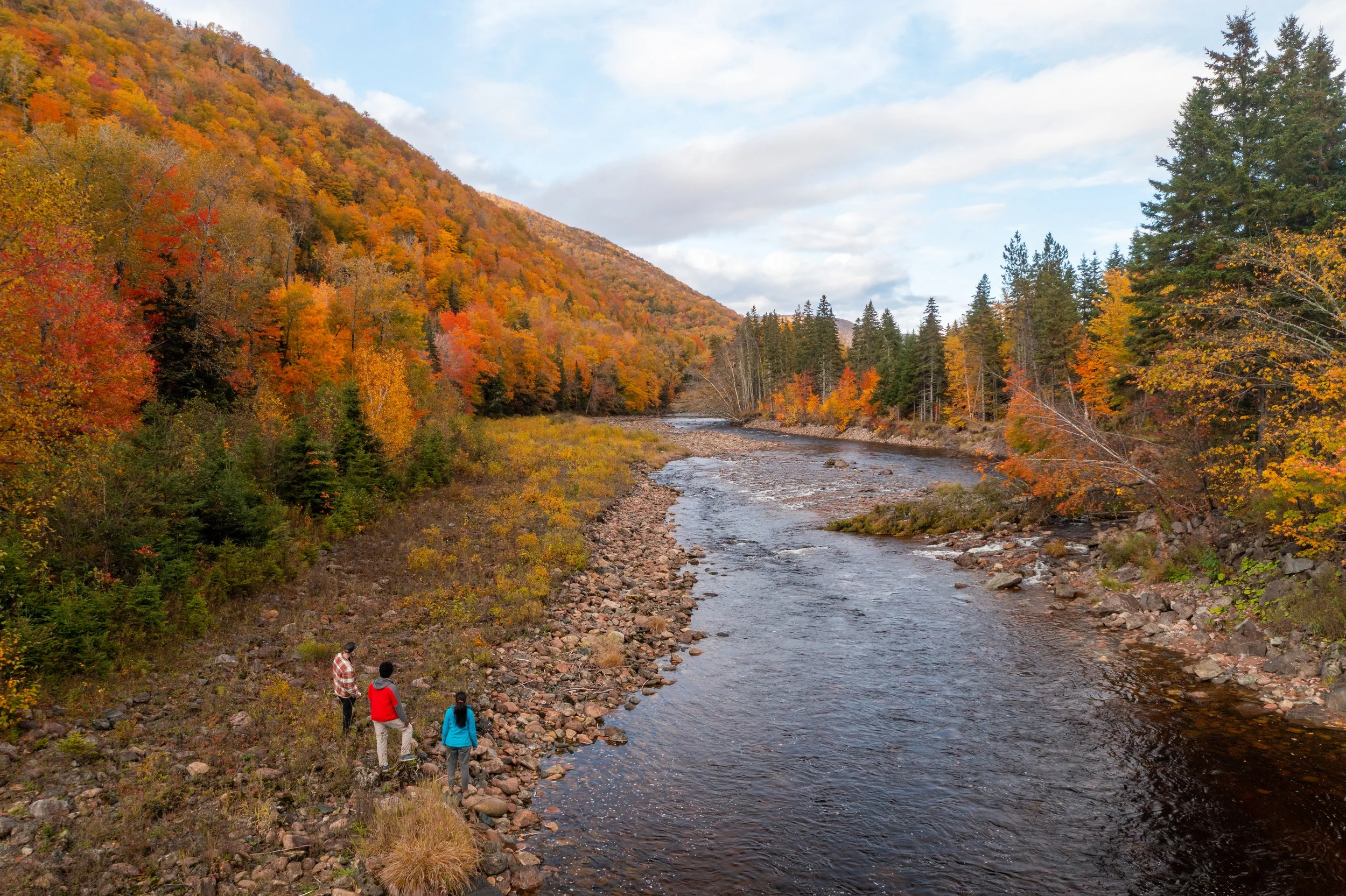 Two people standing beside a river with vibrant fall colours and forested hills in Cape Breton.