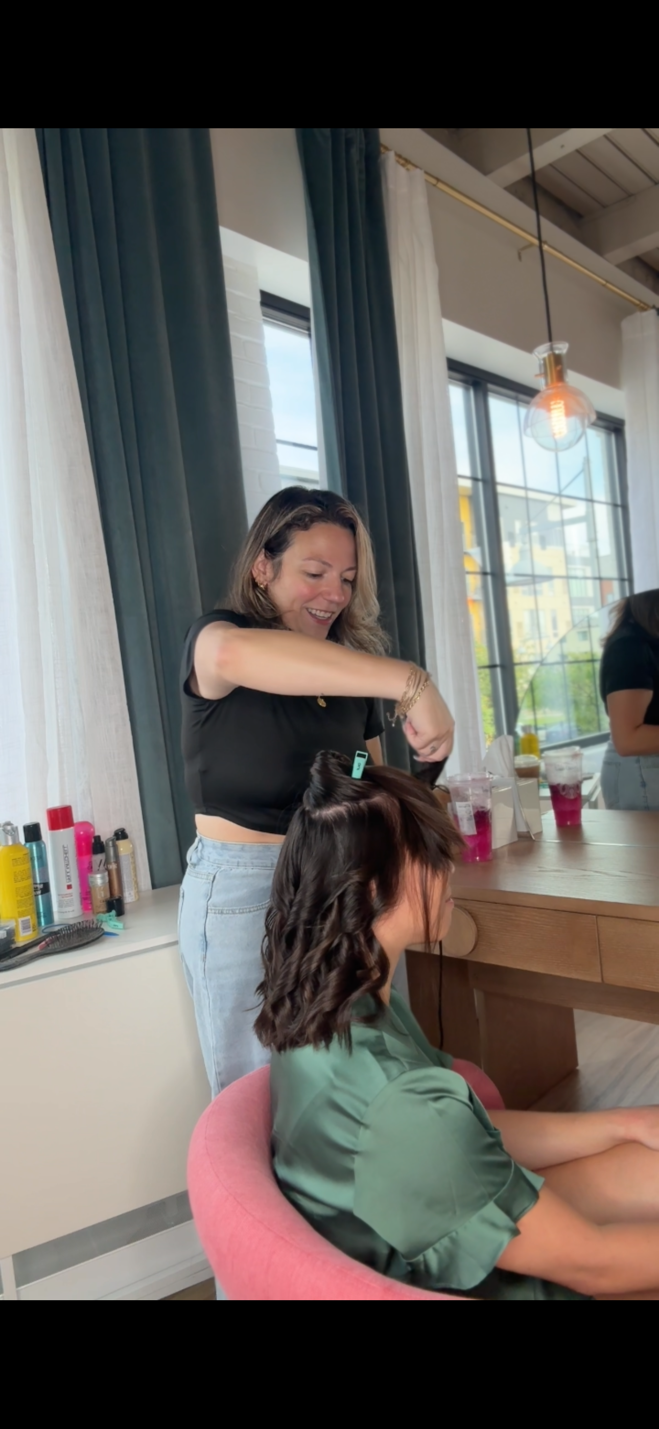A woman is styling another woman's hair in a bright, modern room with large windows, curtains, and a wooden table. The stylist is smiling and working with curling tools, surrounded by hair products and beverages on the table.