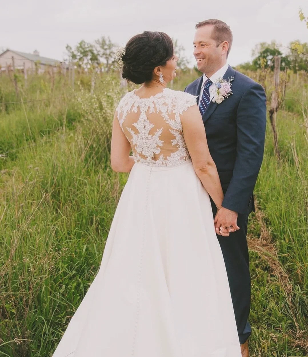 A bride and groom holding hands, facing each other, and smiling outdoors in a grassy field on their wedding day.