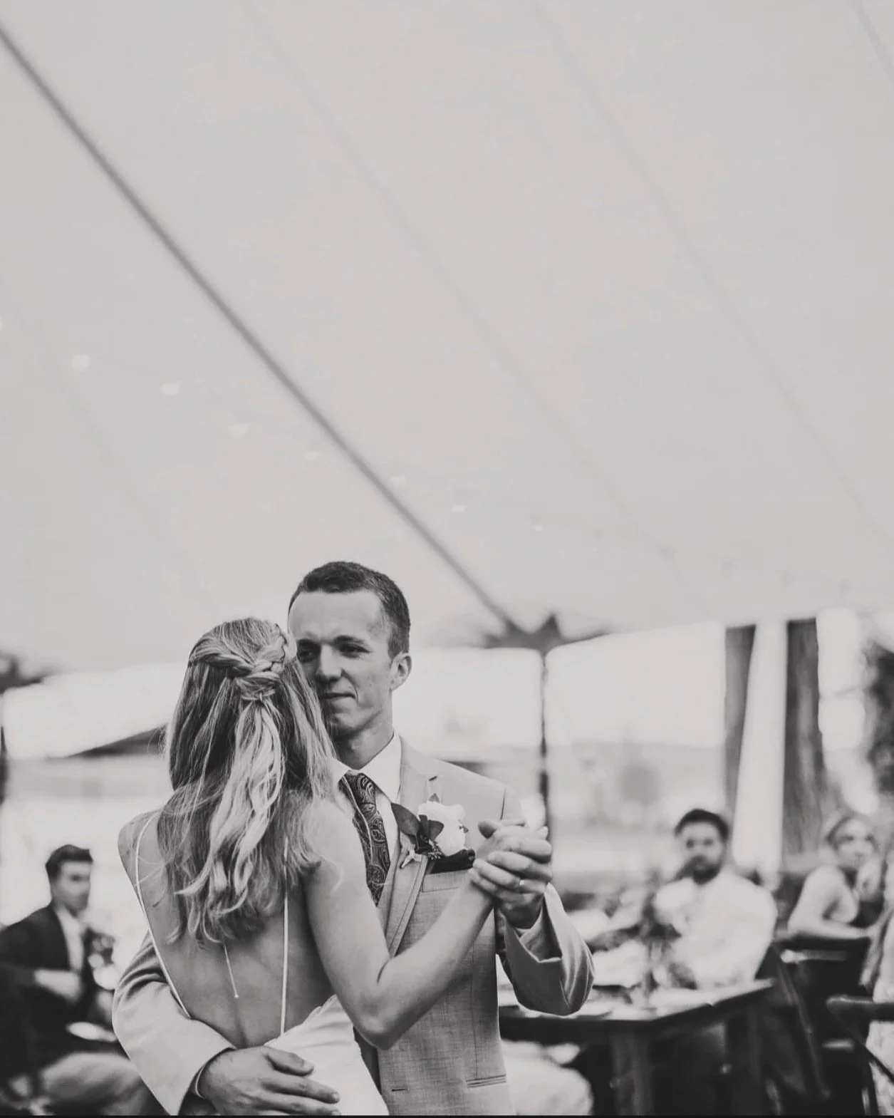 A black and white photo of a wedding couple dancing under a tent, with guests seated at tables in the background.