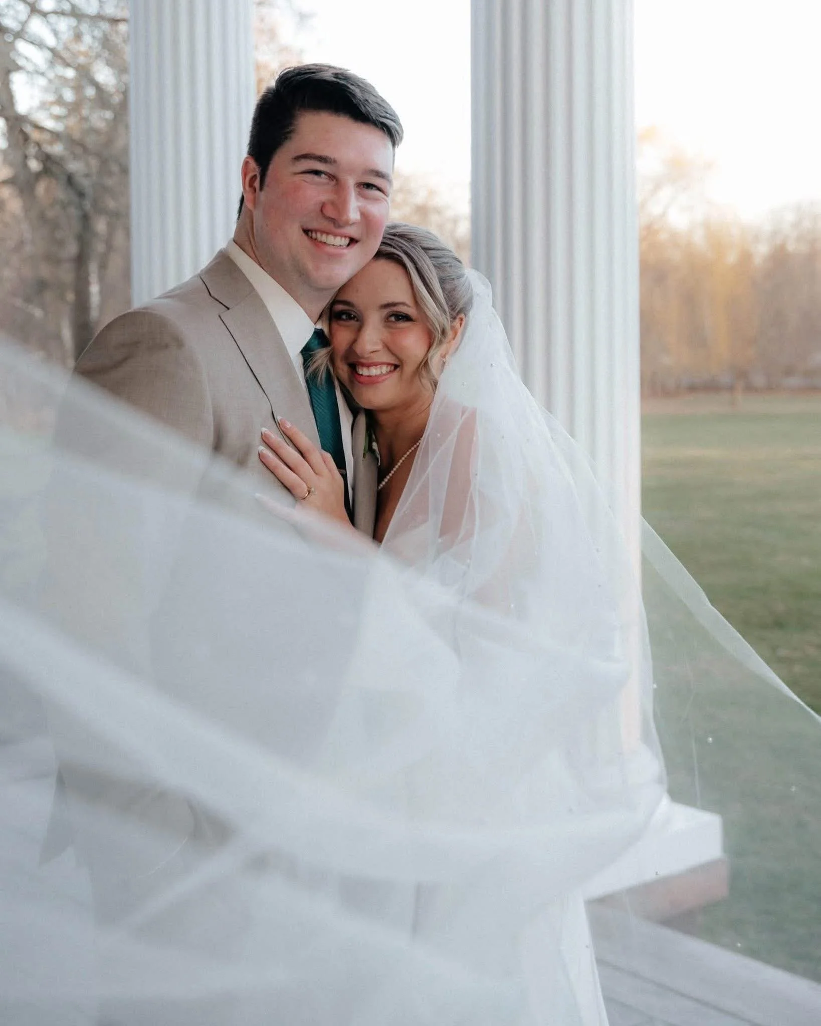 A bride and groom smiling and hugging, standing outside near white pillars and a grassy area with trees.