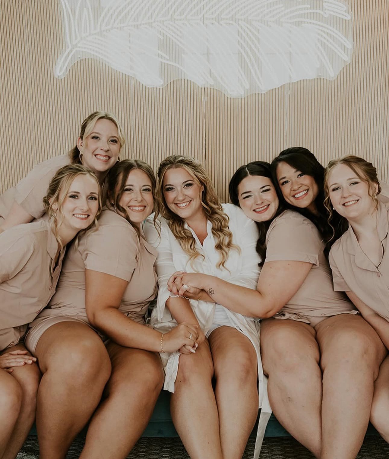 Group of women sitting closely together smiling during a celebration, some wearing matching beige outfits, one in white, in a room with a light wood panel wall and a white decorative cloud arrangement overhead.