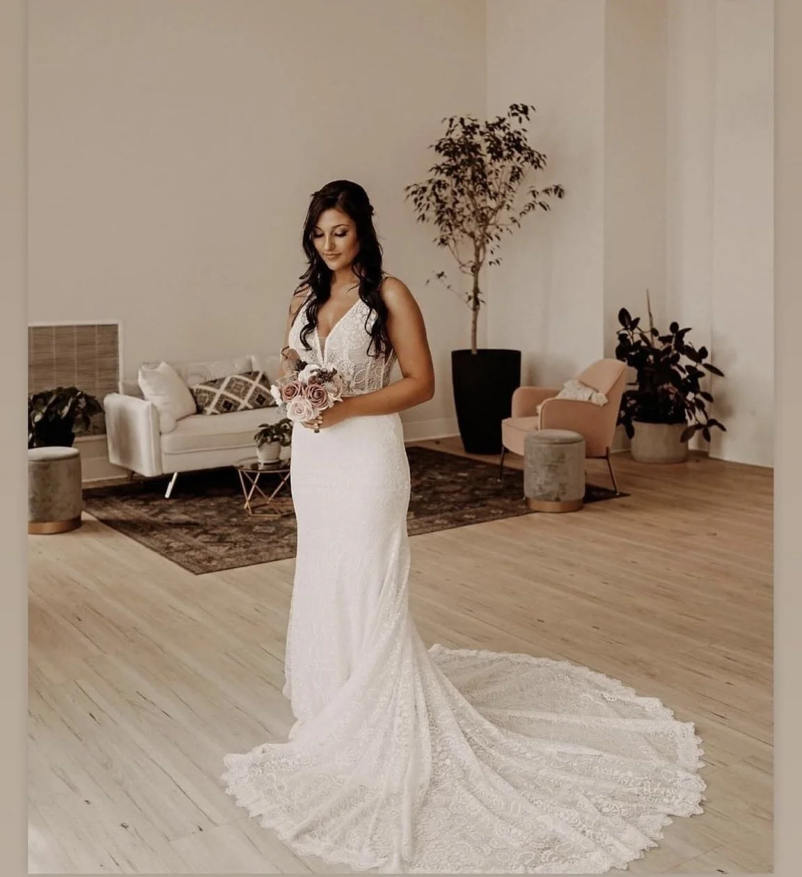 A woman in a white wedding gown holding a bouquet of pink and white roses, standing in a living room with modern furniture and decor.