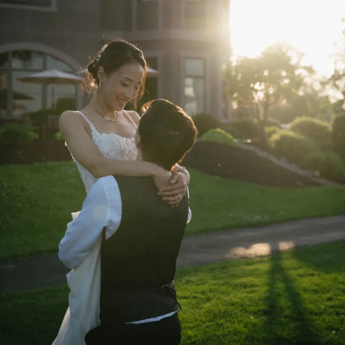 A woman in a white dress being lifted by a man in a vest and shirt during a romantic moment outdoors at sunset, in front of a house with greenery.