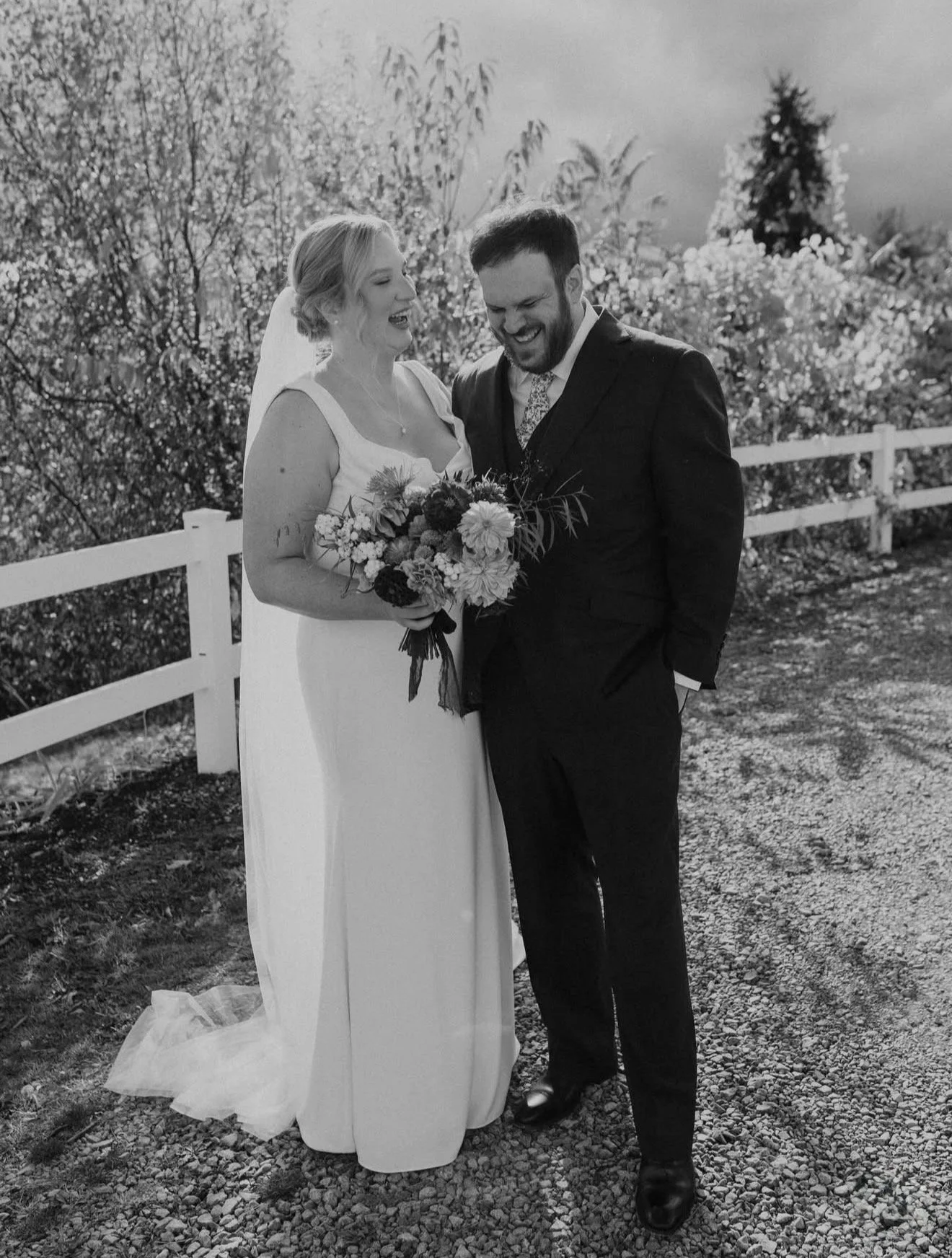 A black and white photo of a bride and groom standing outdoors, smiling and laughing, with the bride holding a bouquet of flowers. The bride is wearing a sleeveless wedding dress and a veil, the groom is in a dark suit and tie, with a white shirt. They are next to a white wooden fence, with trees and cloudy sky in the background.