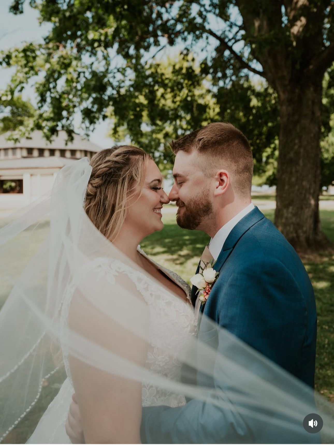 A bride and groom wearing wedding attire, standing close with foreheads touching, smiling outdoors under a large tree during daytime.