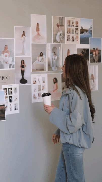 A woman with long brown hair, wearing a light blue jacket and jeans, holding a coffee cup, looking at a wall displaying numerous fashion photographs and images of models.
