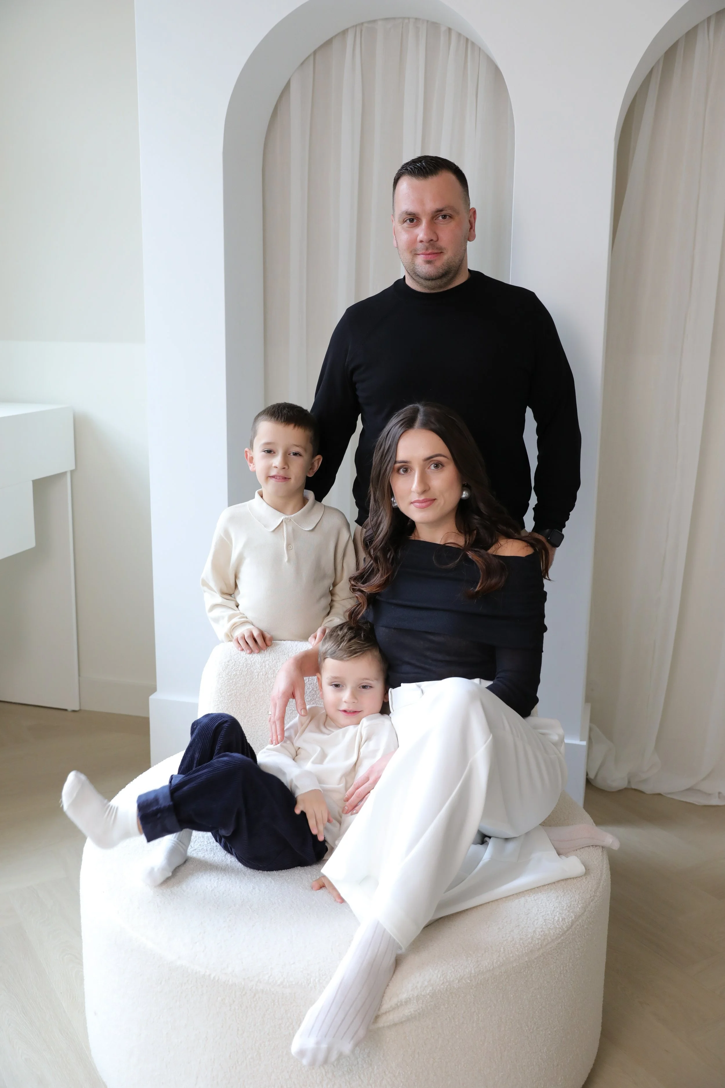 A family portrait featuring a man, woman, and three children in a minimalistic, elegant room with white walls, draped curtains, and a round white upholstered ottoman.