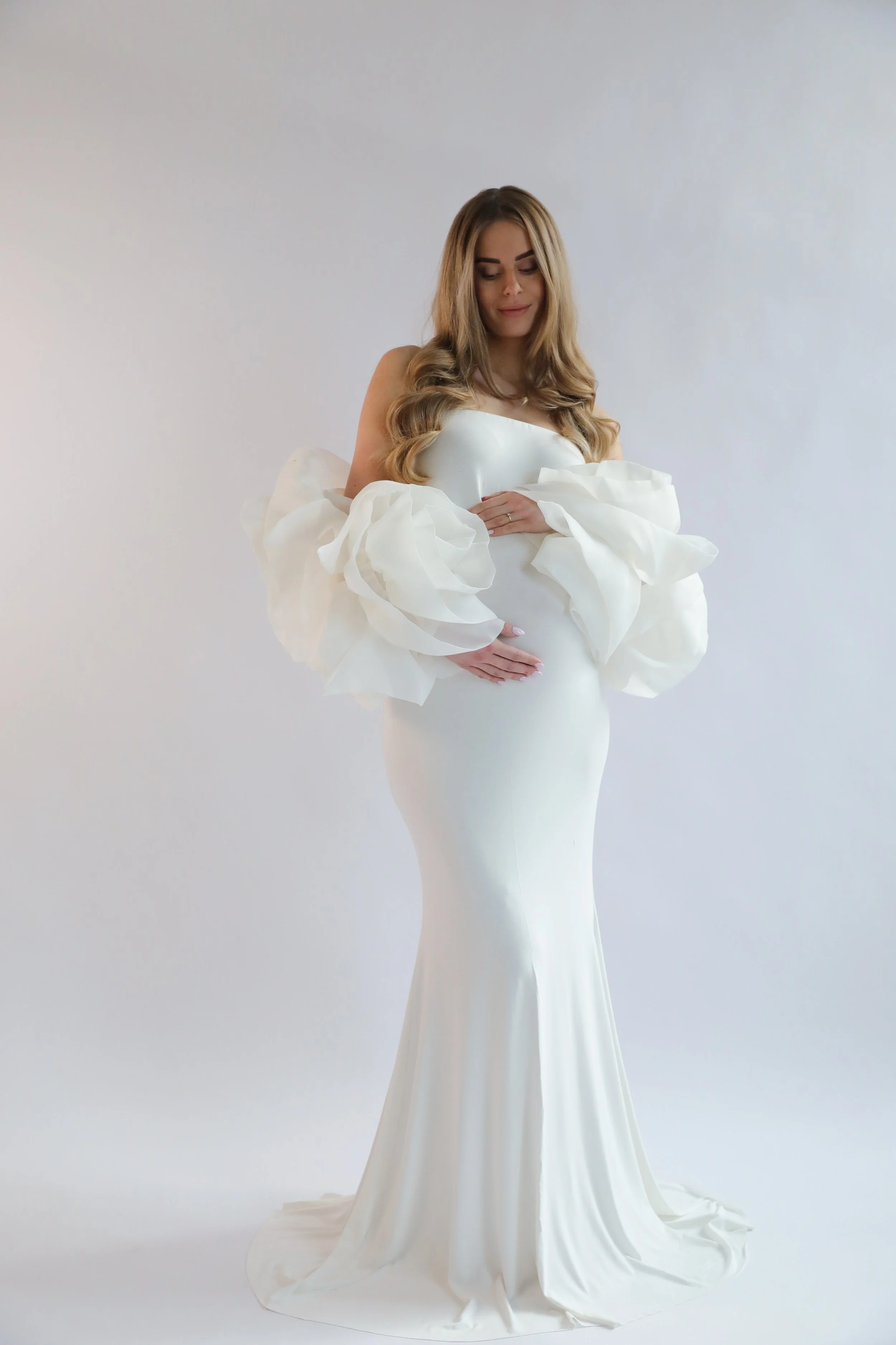 A woman in a white, elegant, strapless gown with large, ruffled sleeves features her standing against a plain background, with long, wavy blonde hair, downward gaze, and gentle expression.