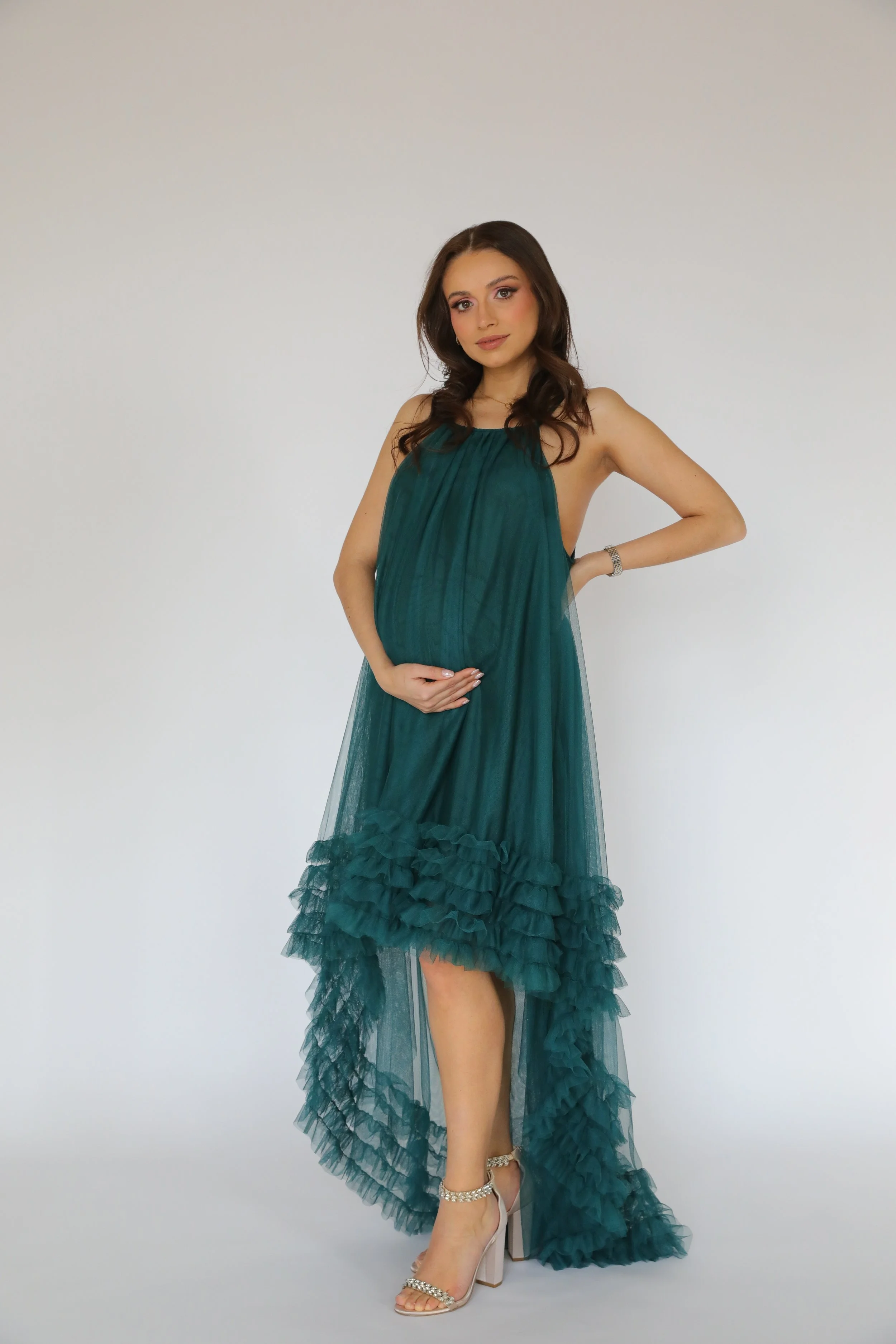A woman wearing a teal evening gown with ruffled layers at the hem, high heels with ankle straps, and a watch, standing against a plain white background.