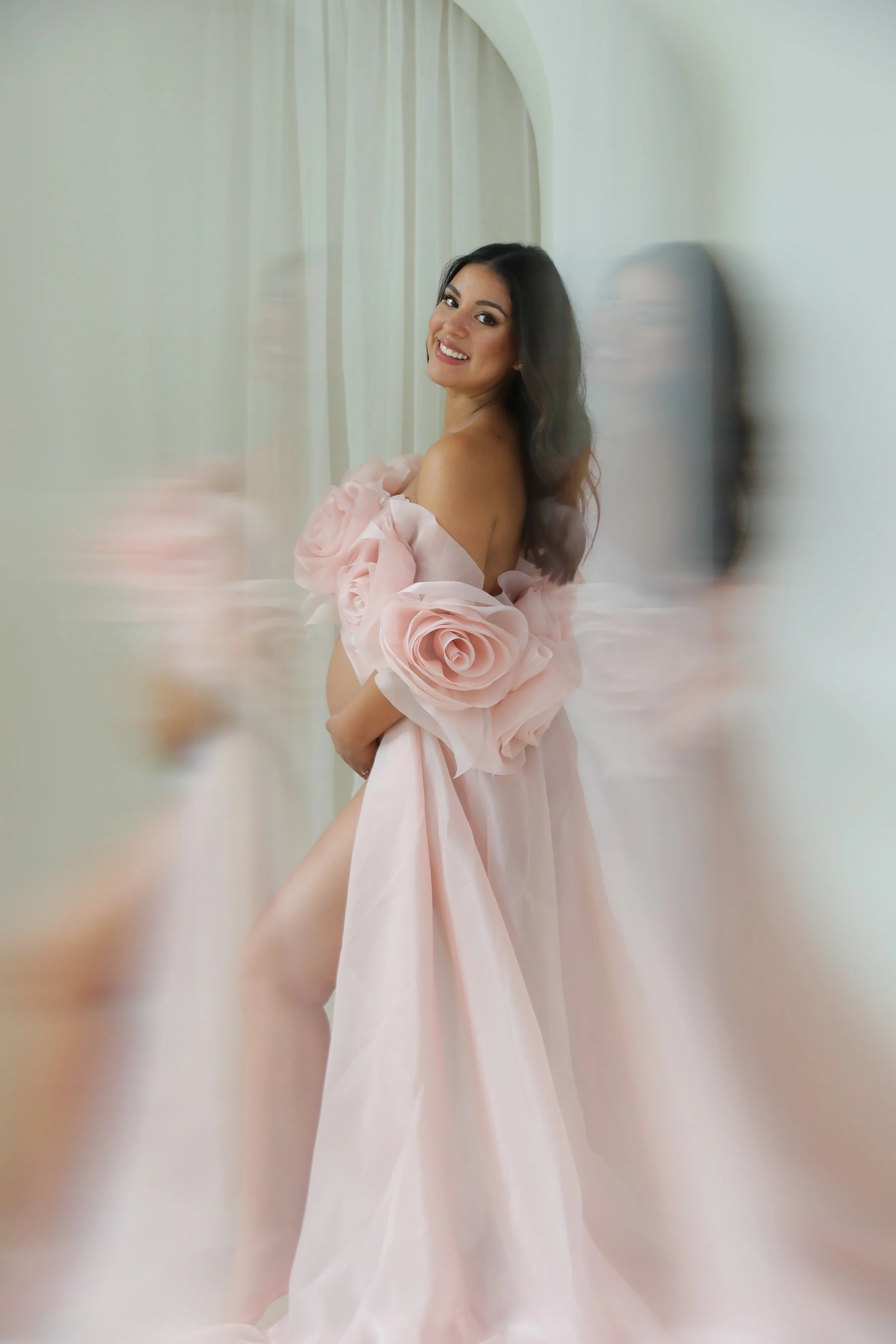 A woman with long dark hair smiling, wearing a pink dress with large floral details on the shoulders, standing in front of a light-colored background with curtains and a curved arch.