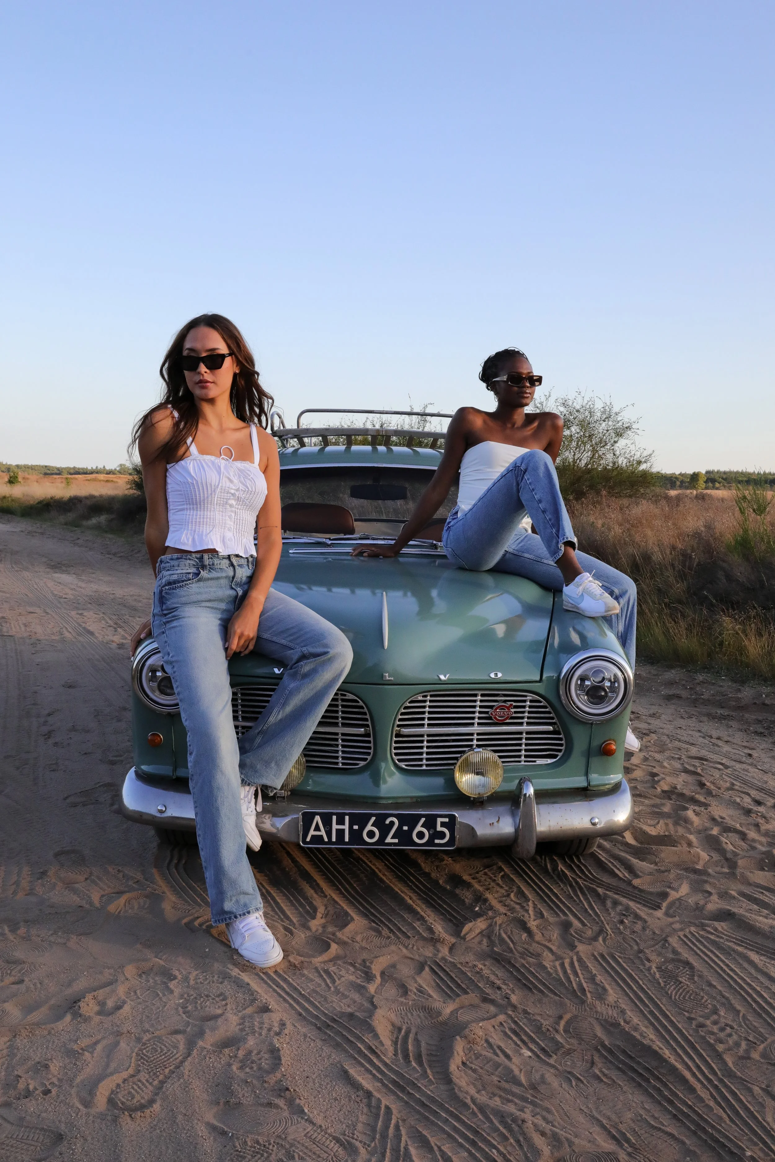 Two women wearing sunglasses and casual clothing pose on a vintage green car on a dirt road in an open rural landscape during sunset.