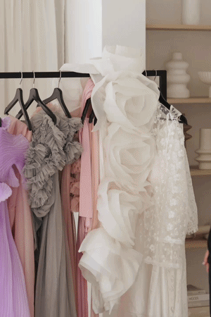 Clothes hanging on a black rack, including dresses in purple, gray, pink, and white with large fabric flower details.