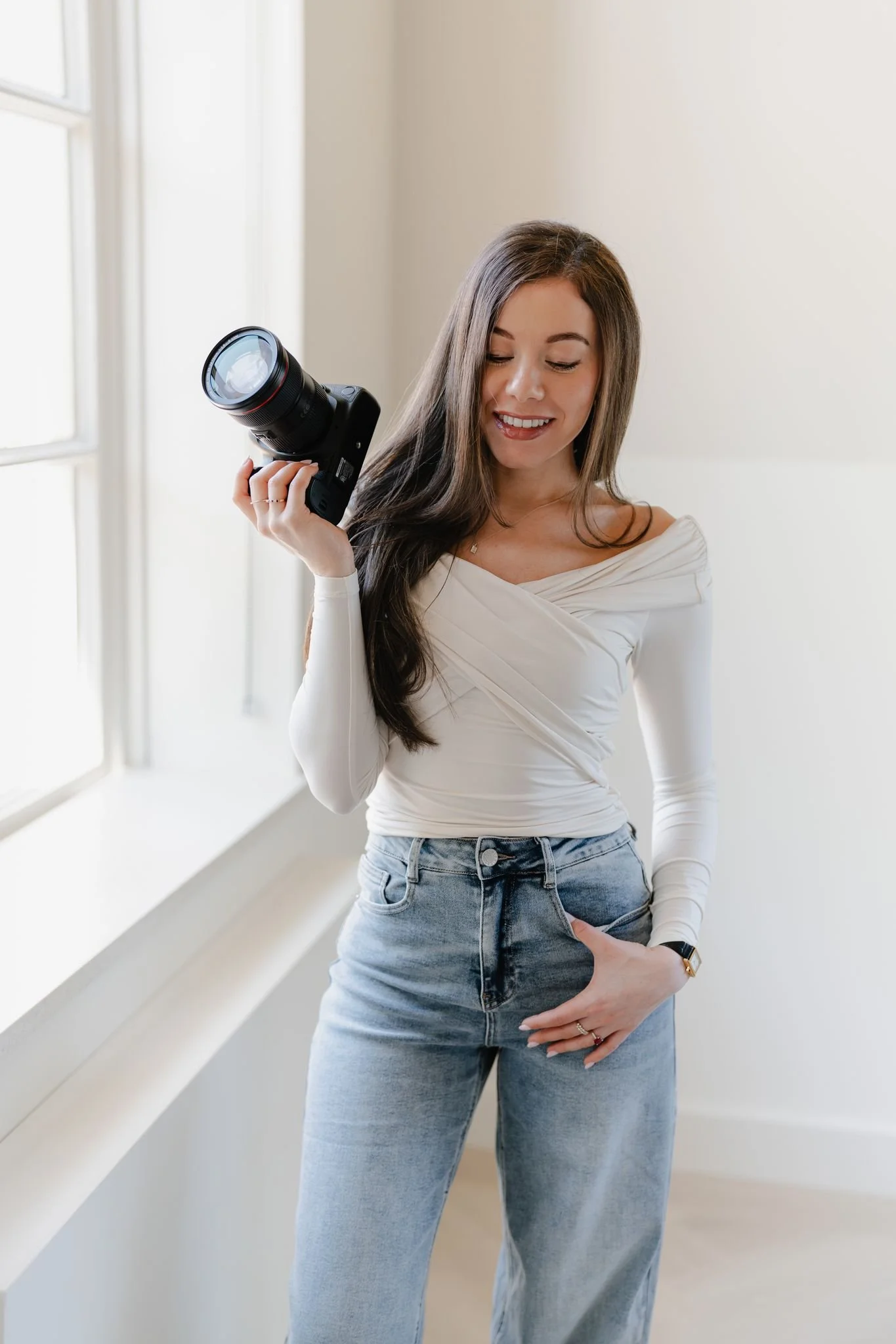 A woman with long brown hair wearing a white off-the-shoulder top and blue jeans, holding a camera and smiling, standing near a window in a bright room.
