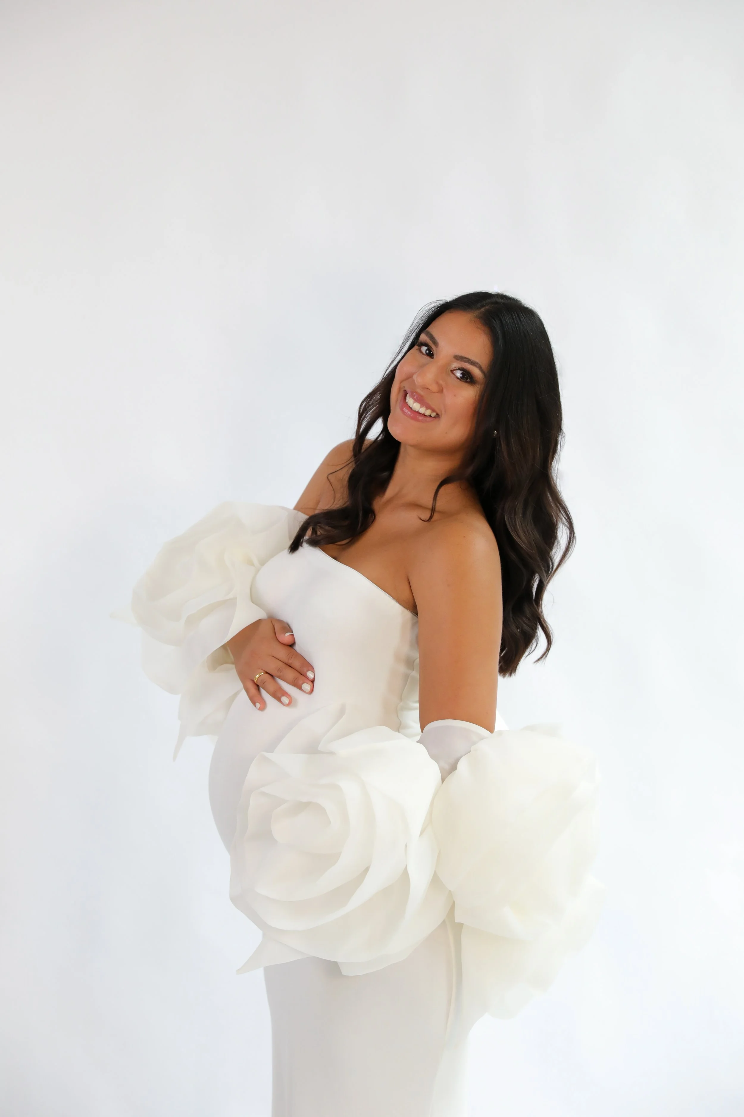 A woman with long dark hair smiling, holding white fabric with large white floral or ruffle details against a plain white background.