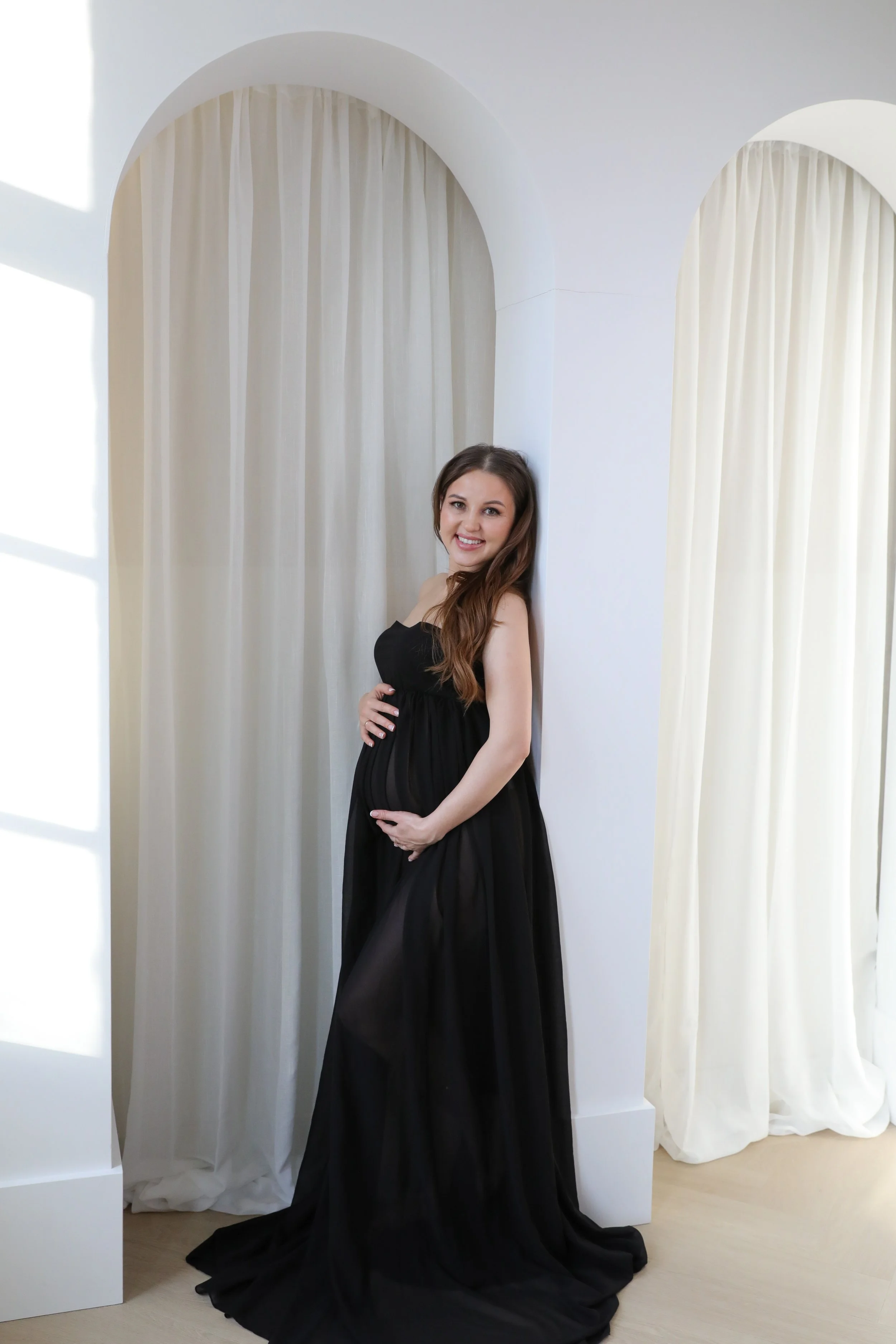 A pregnant woman in a black dress is standing against a white wall and cream-colored curtains, smiling and looking at the camera.