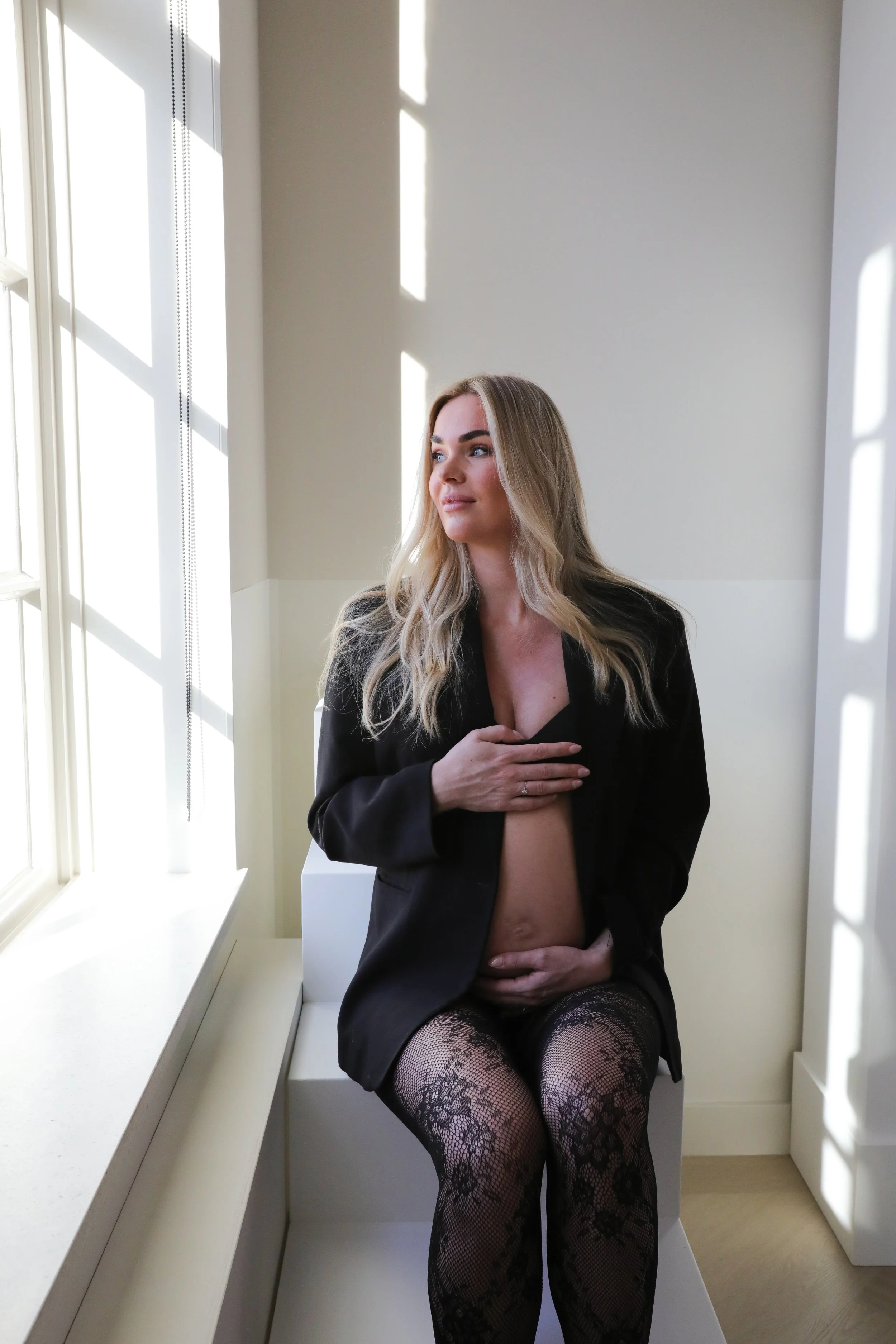A woman with long blonde hair, dressed in black lingerie with fishnet stockings, sits on a white bench by a window with sunlight casting shadow patterns on the wall. She covers her chest with one hand and rests her other hand on her abdomen, looking 
