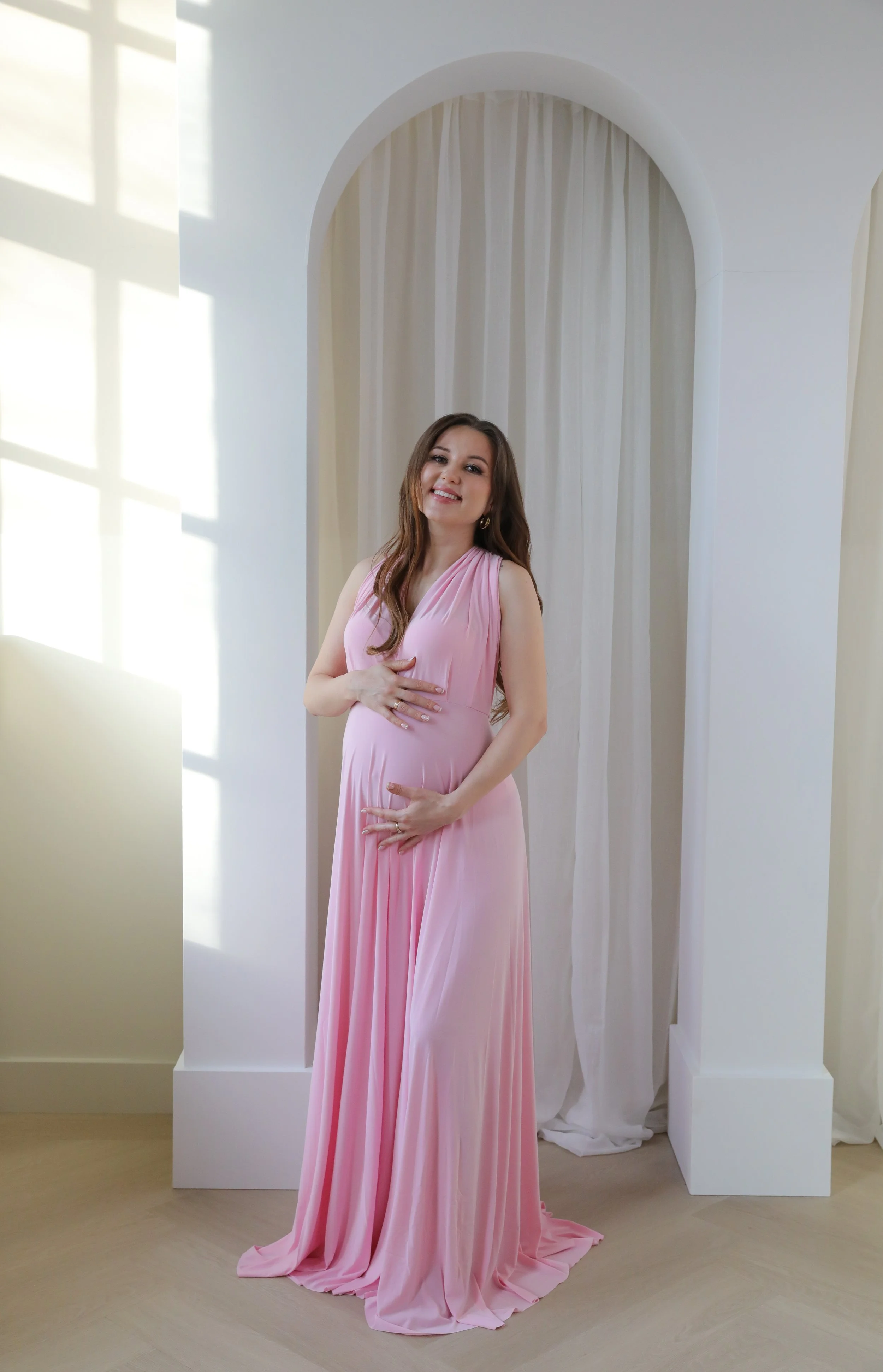 A woman in a long pink gown standing in front of a white archway with curtains, sunlight casting shadow patterns on the wall.