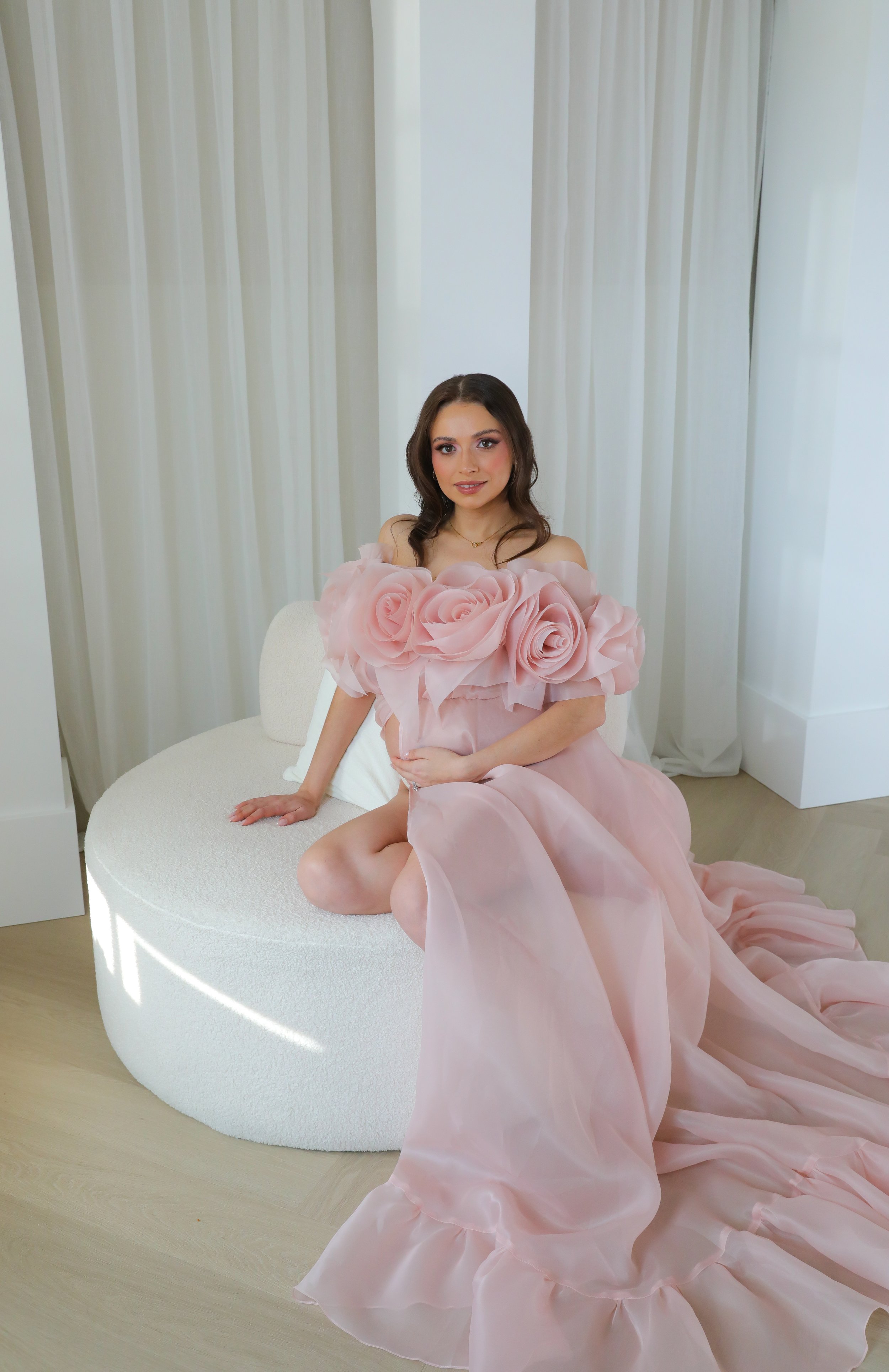 A woman with brown hair, wearing a pink dress with large roses and ruffled fabric, sitting on a white circular bench in a room with white curtains and light wood flooring.