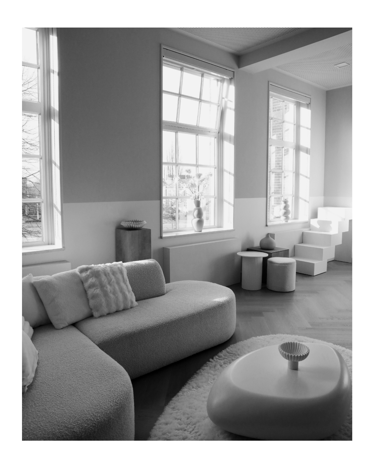 A modern, minimalist living room with large windows, a curved sofa with pillows, a round ottoman, and decorative vases. The room has natural light coming through the windows, with a wooden floor and neutral decor.